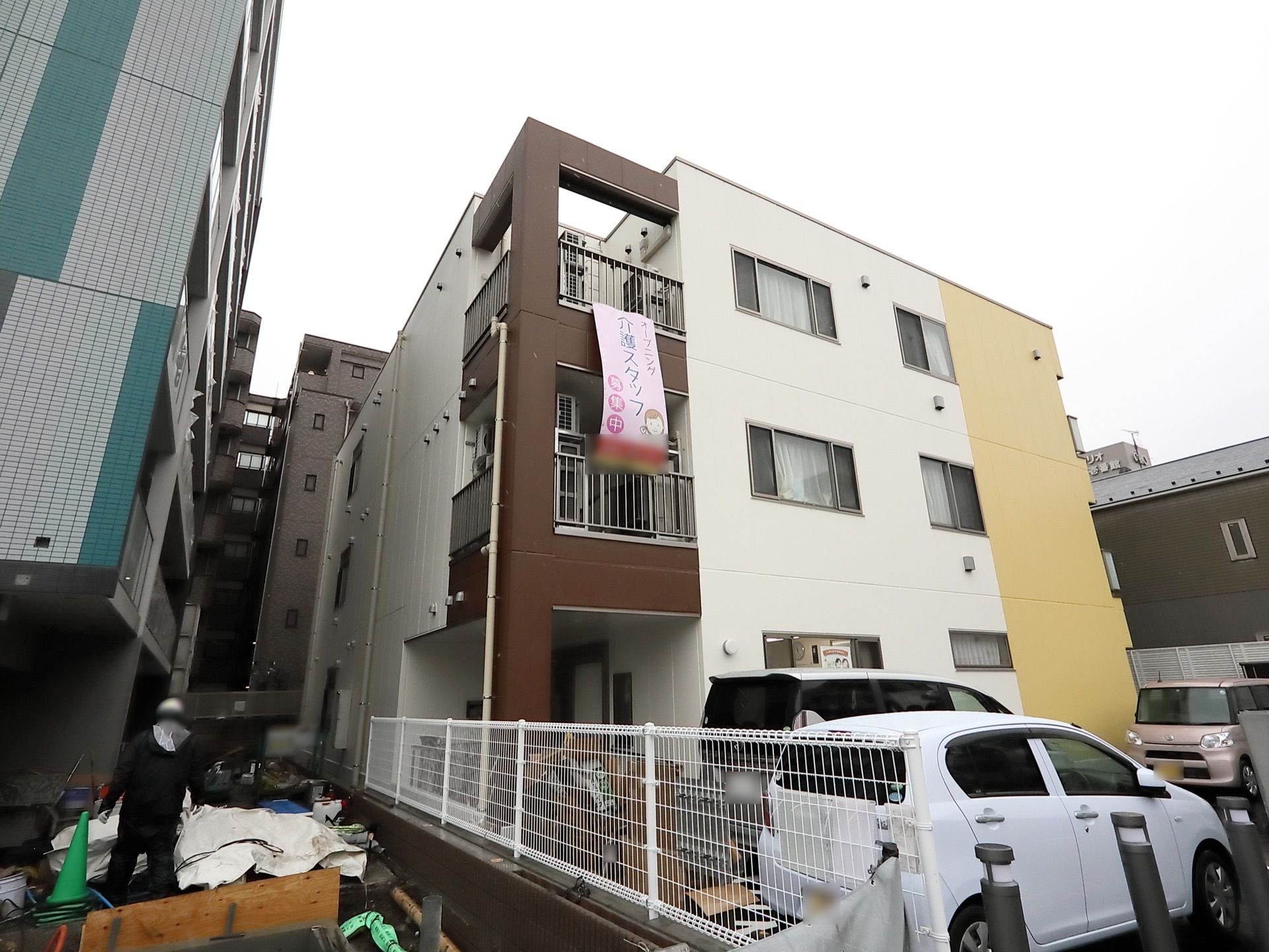 ３階建ての建物で、外壁はブラウンとホワイト、イエローに色分けされている。介護スタッフ募集の垂れ幕が掛かっている。