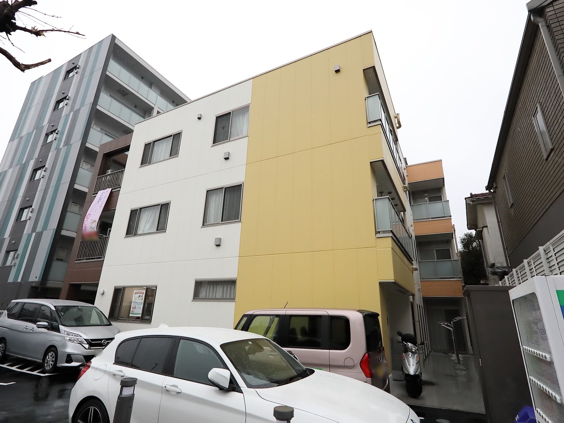 駐車場に白い車が停まっていて、その前に自動販売機があるのが見える。施設の周囲にはマンションや住宅が建ち並んでいる。