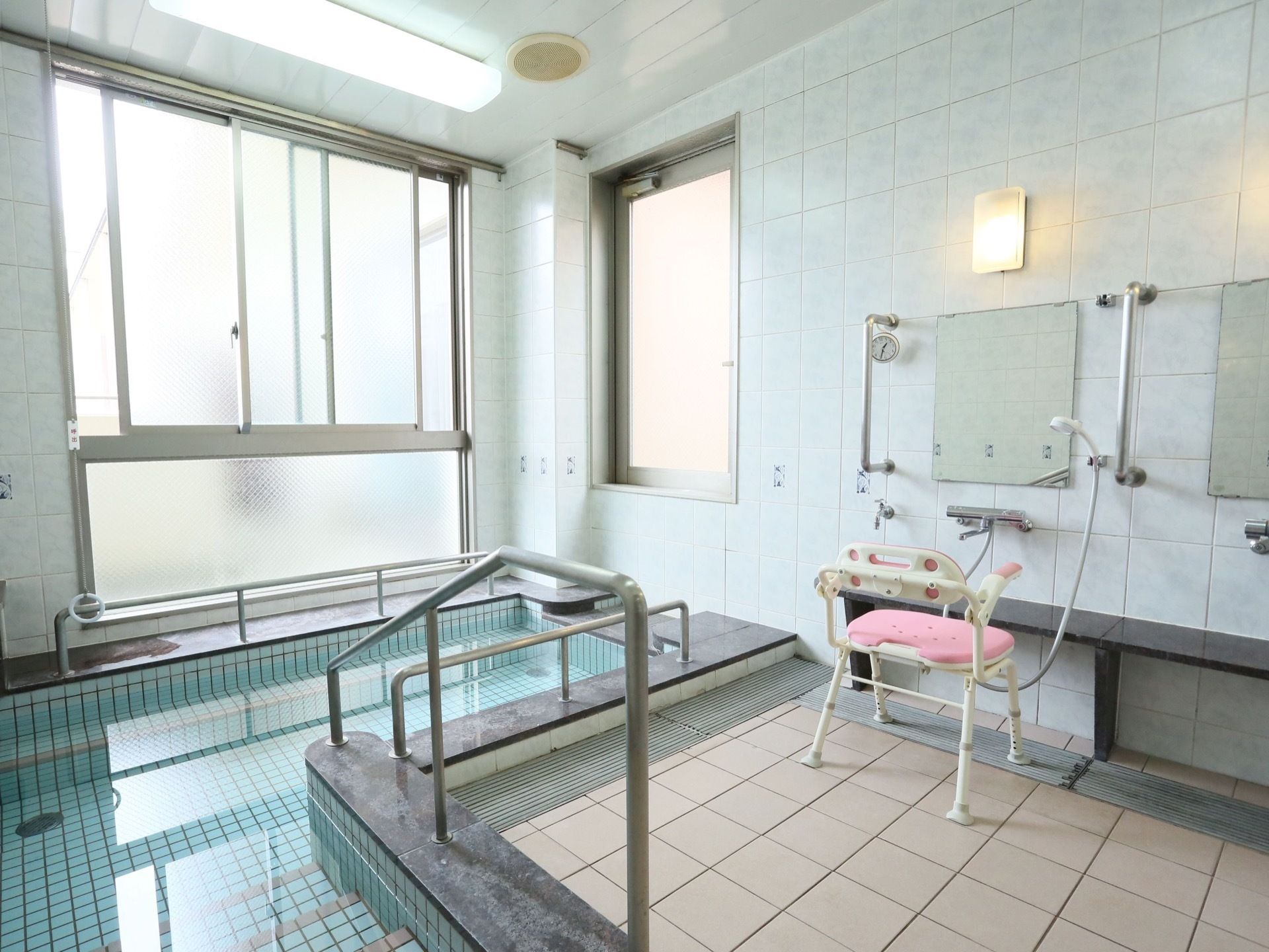 天井まで続く大きな窓のある明るく広い浴室。水色のタイルの壁で囲まれ、大きな浴槽には手すりが設置されている。