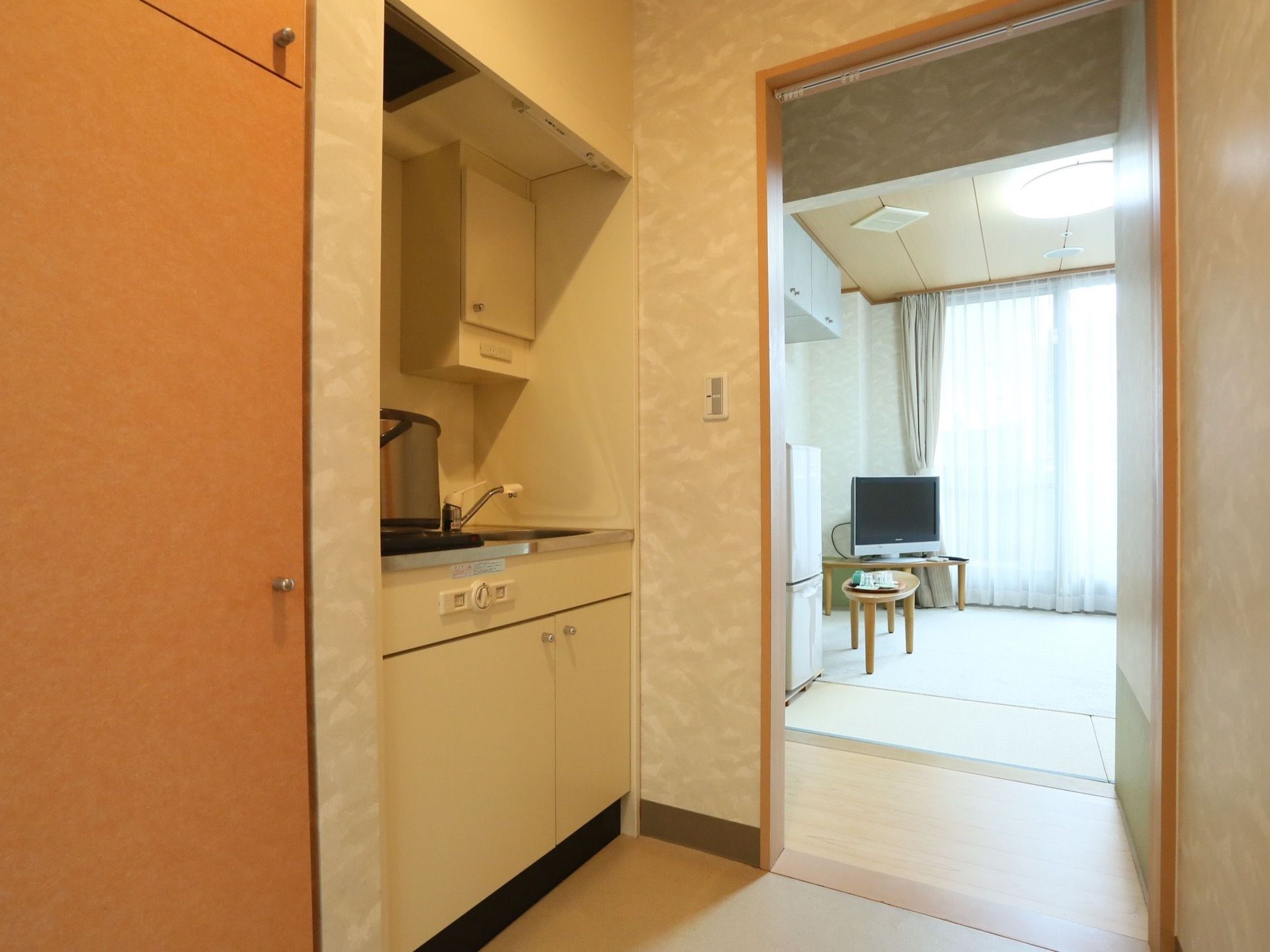 大きな窓のある明るい居室。冷蔵庫、テーブル、テレビが置かれている。入口付近にはミニキッチンが備わっている。