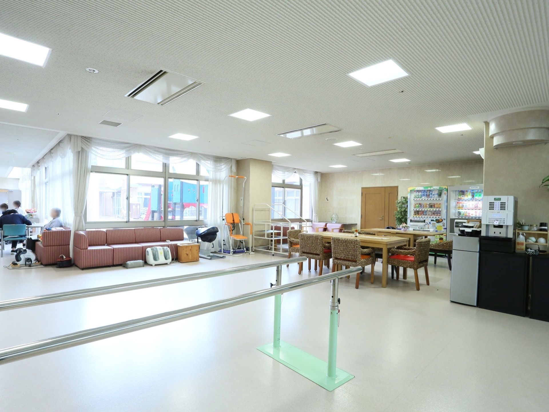 壁前面が大きな窓になっている広い談話室がある。歩行練習用の手すりが置かれ、窓際にはソファがある。自動販売機が置かれている。