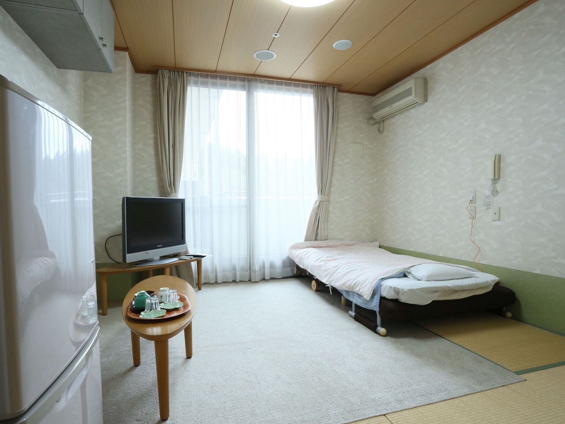 大きな窓のある明るい居室。ベッド、テレビ、冷蔵庫、テーブルが置かれている。天井付近には収納棚がある。畳の上にカーペットが敷かれている。