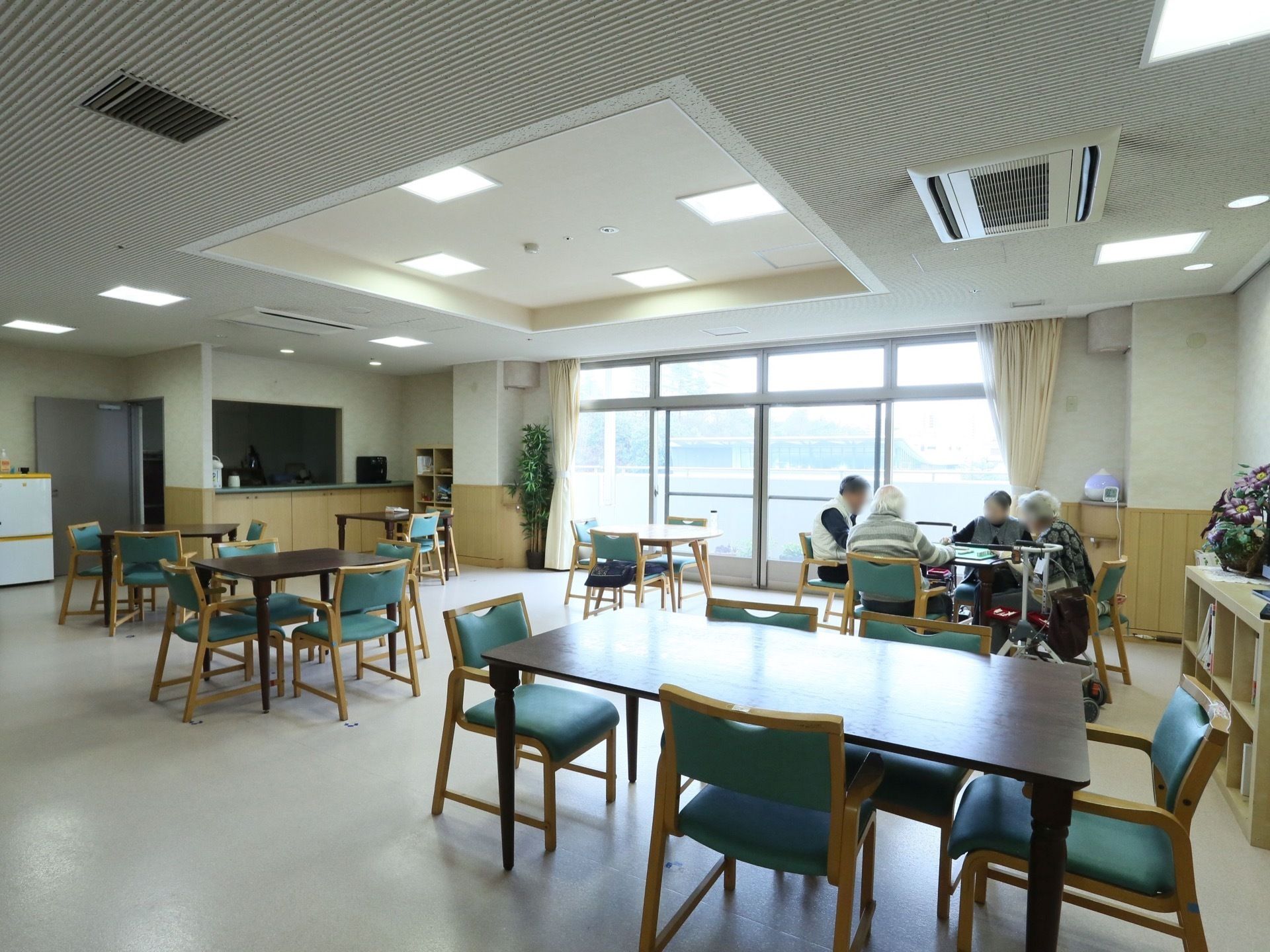 壁一面が大きな窓になっている明るい食堂。テーブルと椅子がたくさん並べられている。植木鉢と本棚が置かれている。