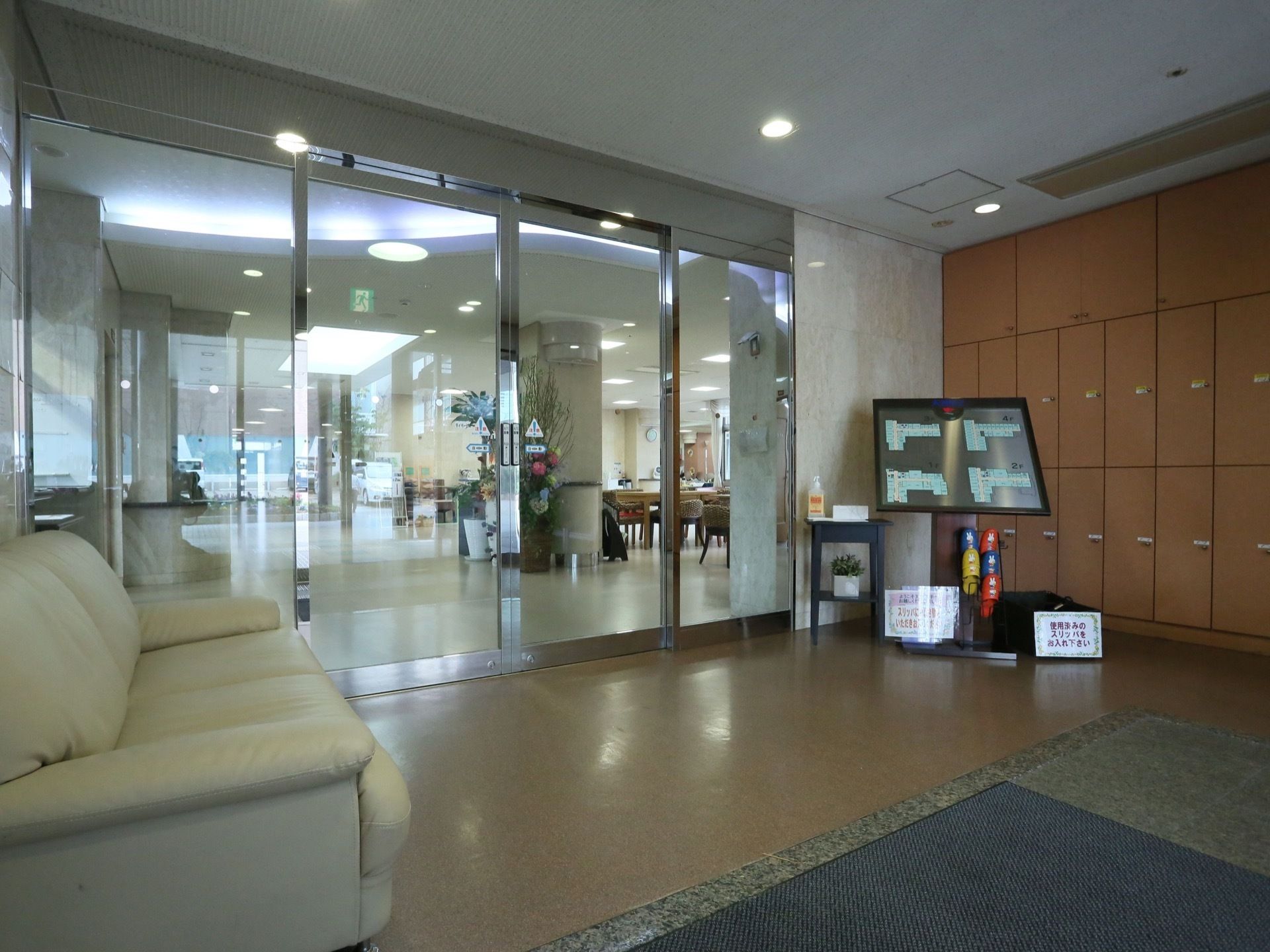玄関はガラス張りの自動ドアで、光沢のある金属フレームを採用している。壁には収納棚があり、大きなソファと施設内の案内図が置かれている。