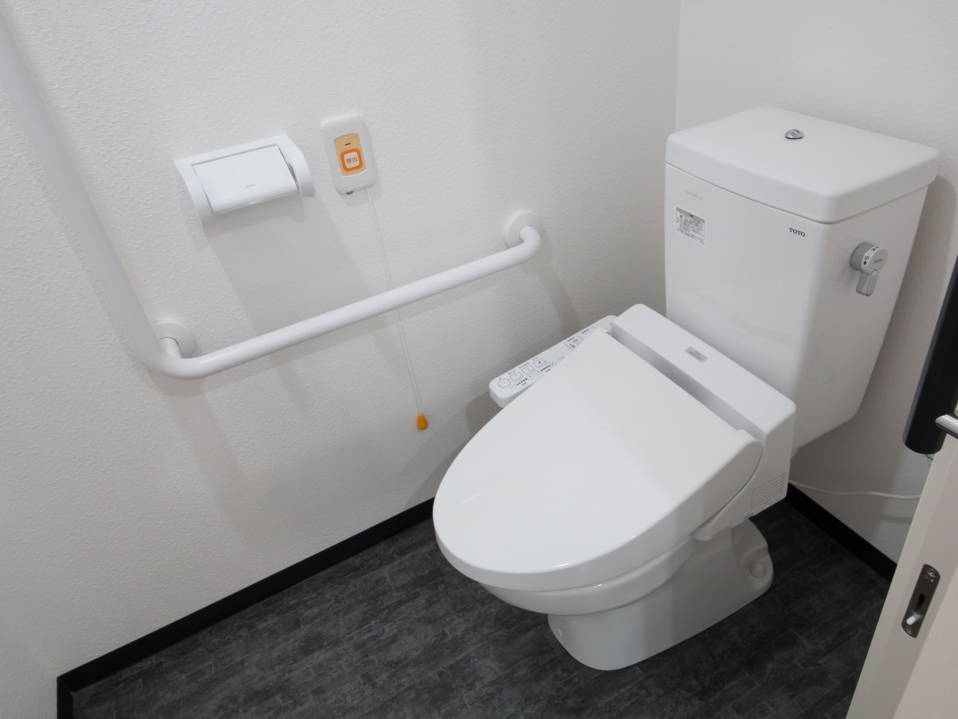 居室のトイレである。壁にはL字型の手すりがあり、長い紐が付属している緊急時呼び出しボタンが設置されている。