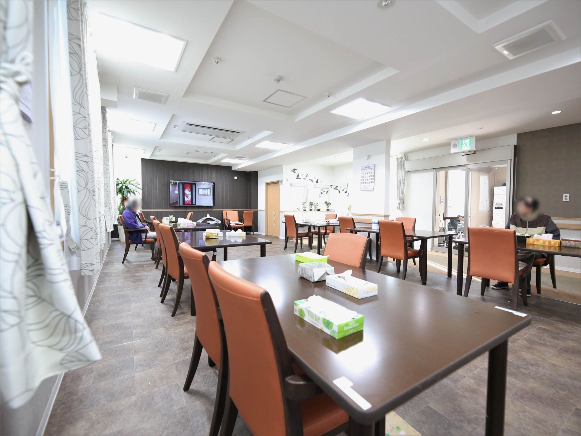 広い食堂にテーブルと赤茶色の椅子が2列並んでいる。カーテンの掛かった窓があり、床と壁の一部がダークブラウンになっている。