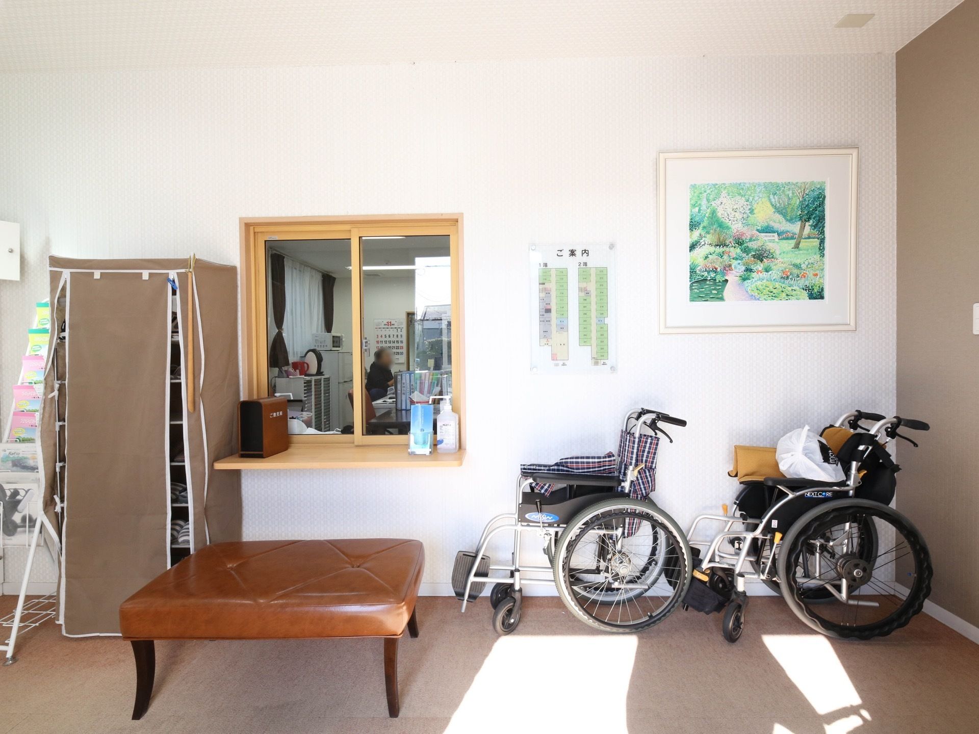 玄関を入った先にあるホール。ガラス窓のある受付窓口があり、手前にはクッションの貼られたベンチが置かれている。壁には絵が飾られている。