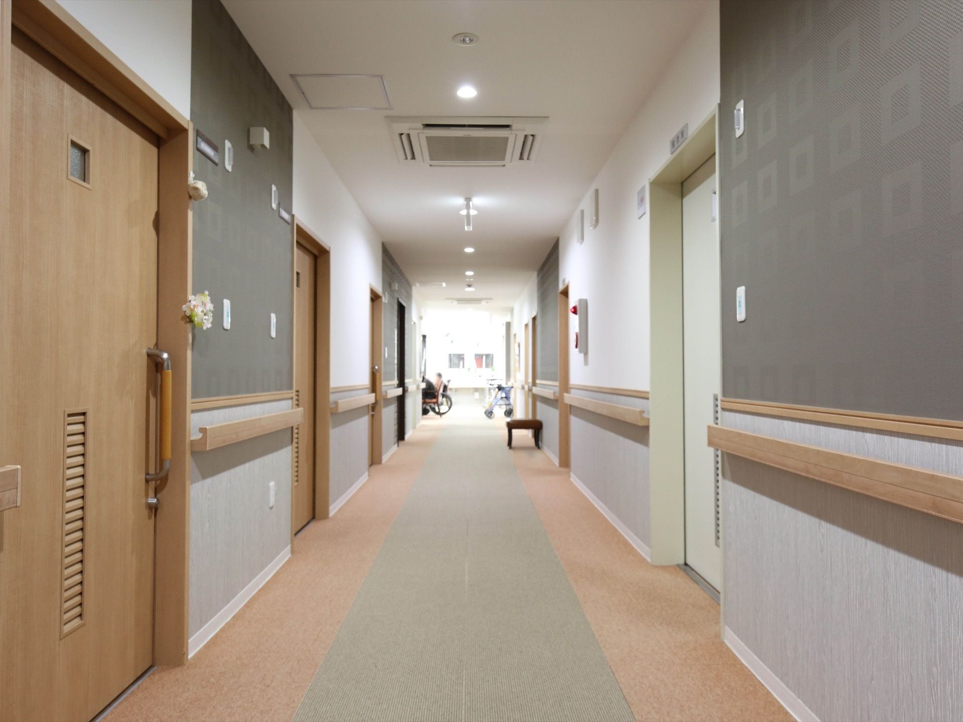 一部がグレーに塗られた壁の廊下がある。両サイドには居室のドアが並んでいる。壁には手すりが設置されている。