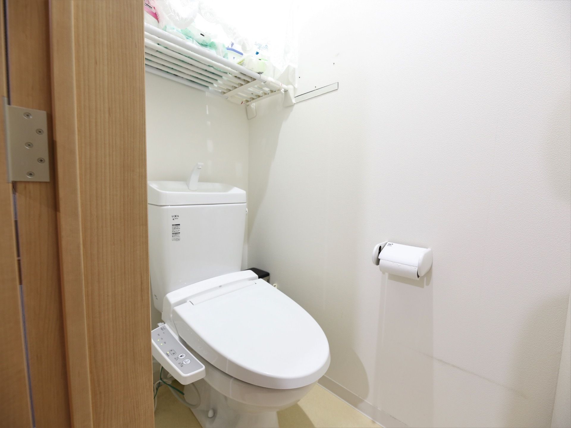 白い壁に囲まれた個室のトイレがある。便器には蓋と温水洗浄便座が付いている。天井付近には突っ張り棚があり、物が置かれている。