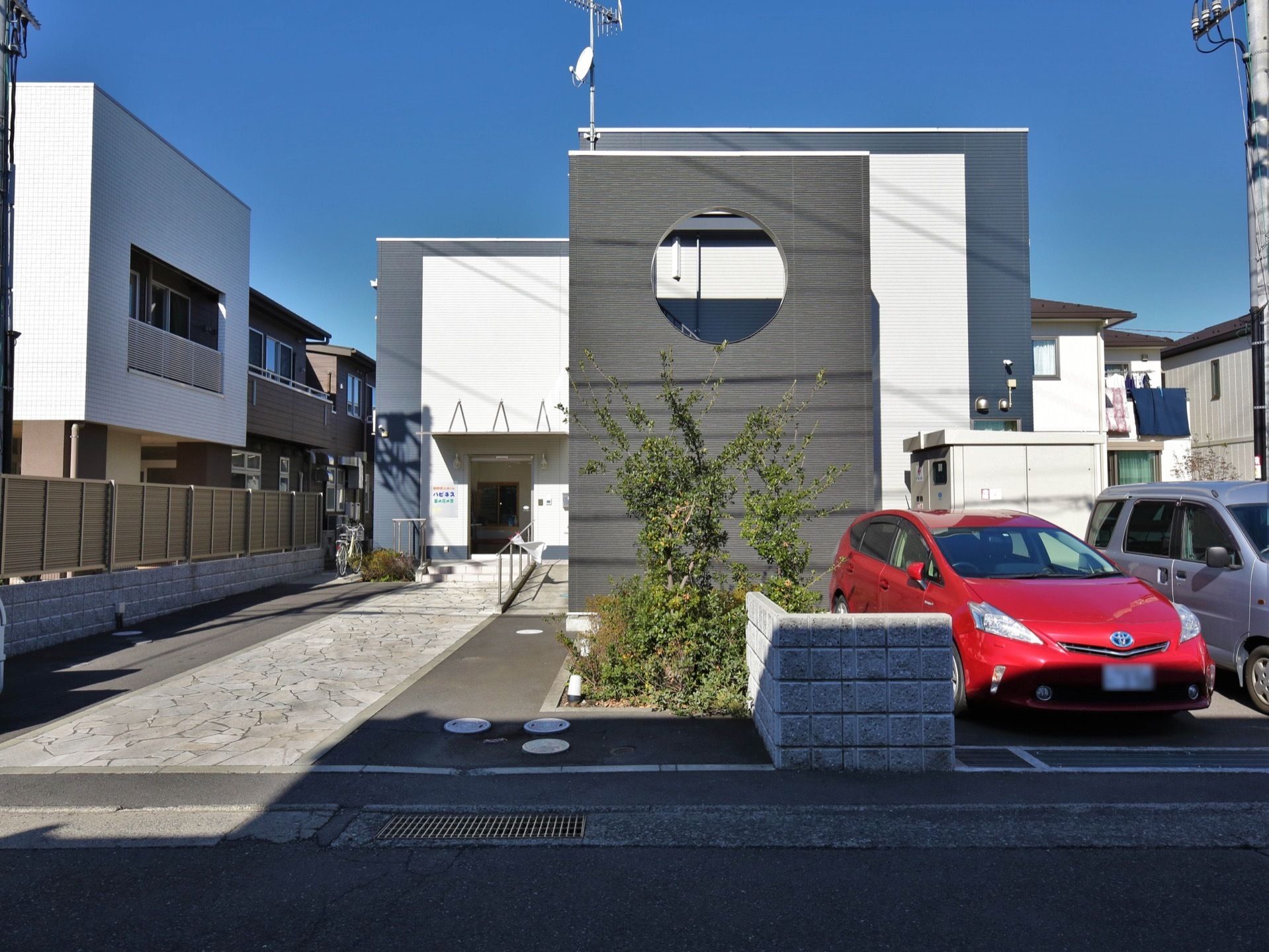 白とグレーを基調としたツートンカラーのモダンなデザインの建物がある。入り口付近には植え込みがあり、隣のスペースは駐車場になっている。