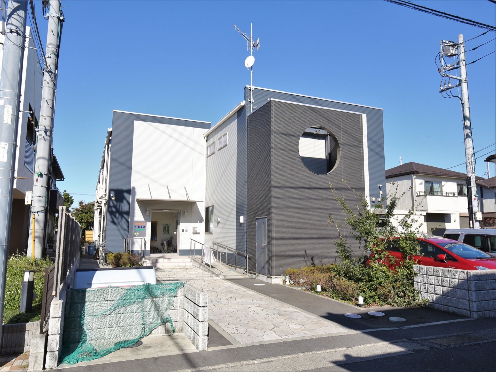 白とグレーの外壁の２階建ての建物がある。玄関までの通路は舗装され、石造り風のタイルが敷き詰められている。手前には緑が繁っている。