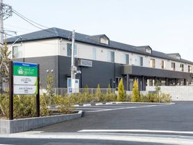 駐車場が付いたベージュの建物。玄関には屋根が付き、住宅と併設介護事業所の看板もある。