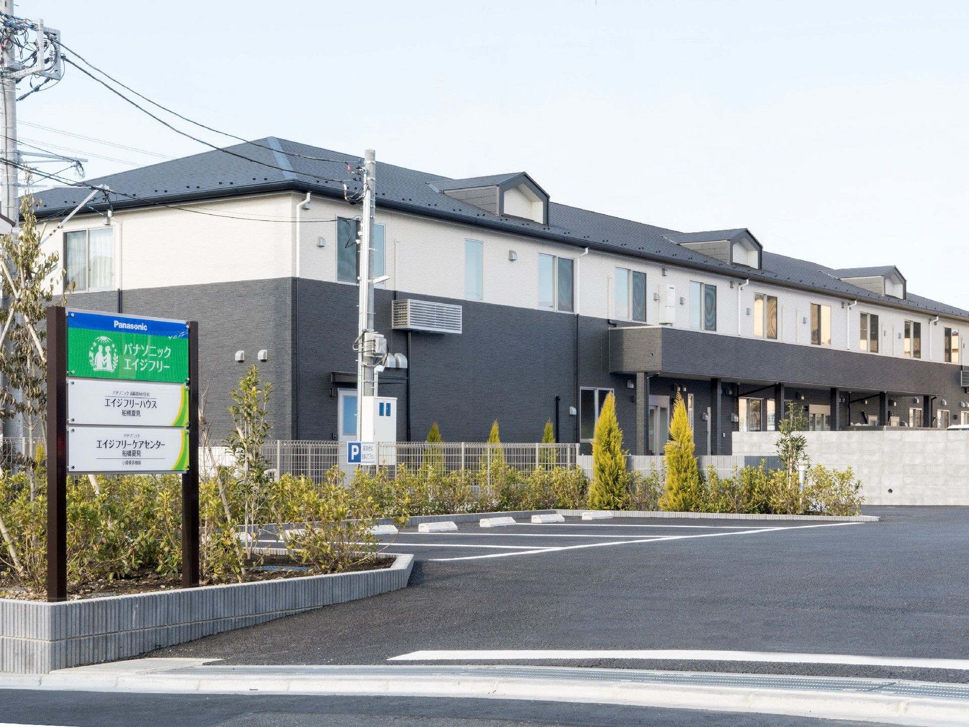 ダークグレーと白の2階建ての建物。建物周辺には木が植えられ、駐車場も用意されている。