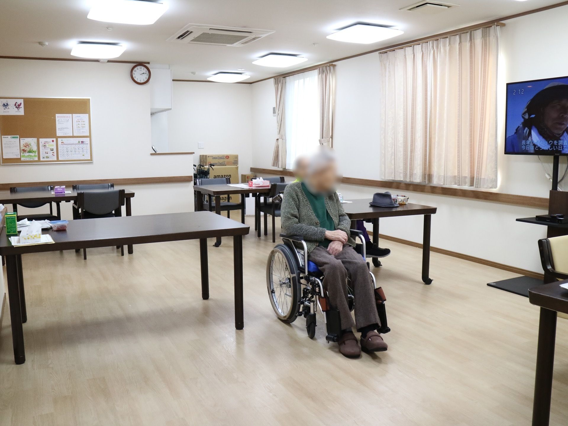 テーブルセットが並べられたフロアがあり、壁際に大型テレビが設置されている。車椅子の入居者様が、1人でテレビを観ている。