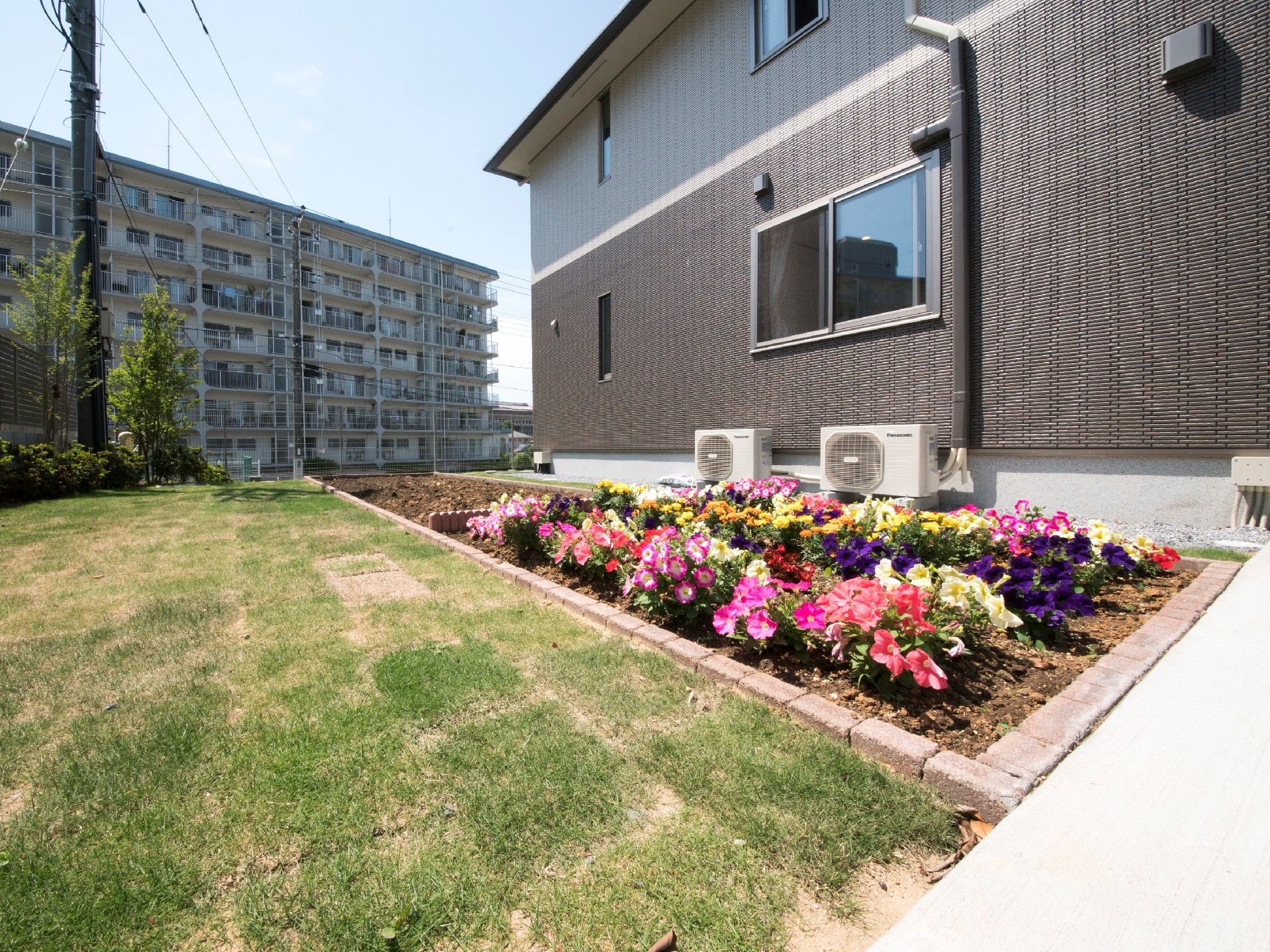 2階建ての建物の前には芝生スペースと花壇があり、奥には大きな集合住宅のような建物が見える。