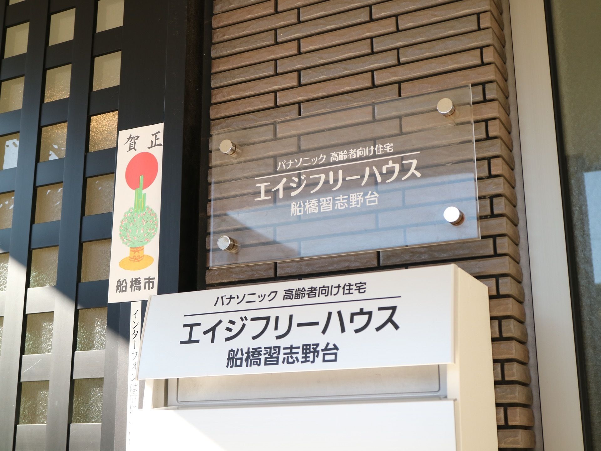 施設扉横のレンガ調外壁に施設プレートが設置されている。透明アクリルボードに白文字で施設名称などが明記されている。