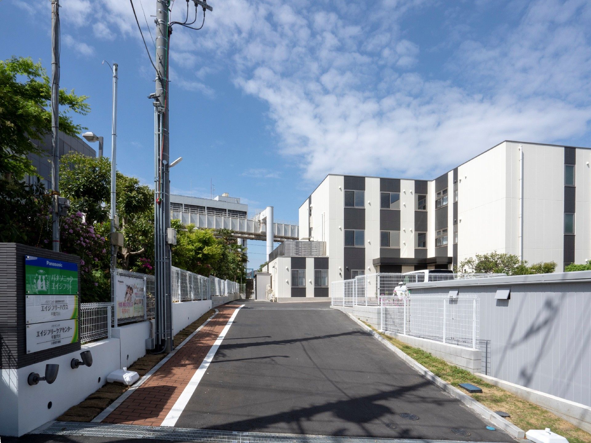 道路の左側には住宅と併設事業所の看板があり、右奥には白い3階建ての建物が見える。