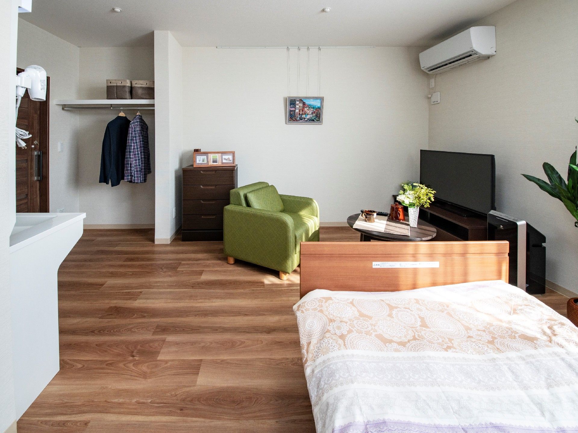 寝具がセットされたベッドが置かれたフローリングの部屋には、テレビやテーブル、エアコンや洗面所もある。