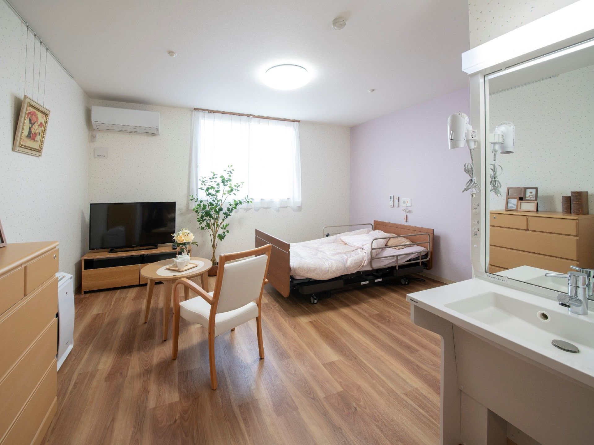 右手には洗面所、正面にはテーブルセットやテレビ、右奥には介護用ベッドがある。
