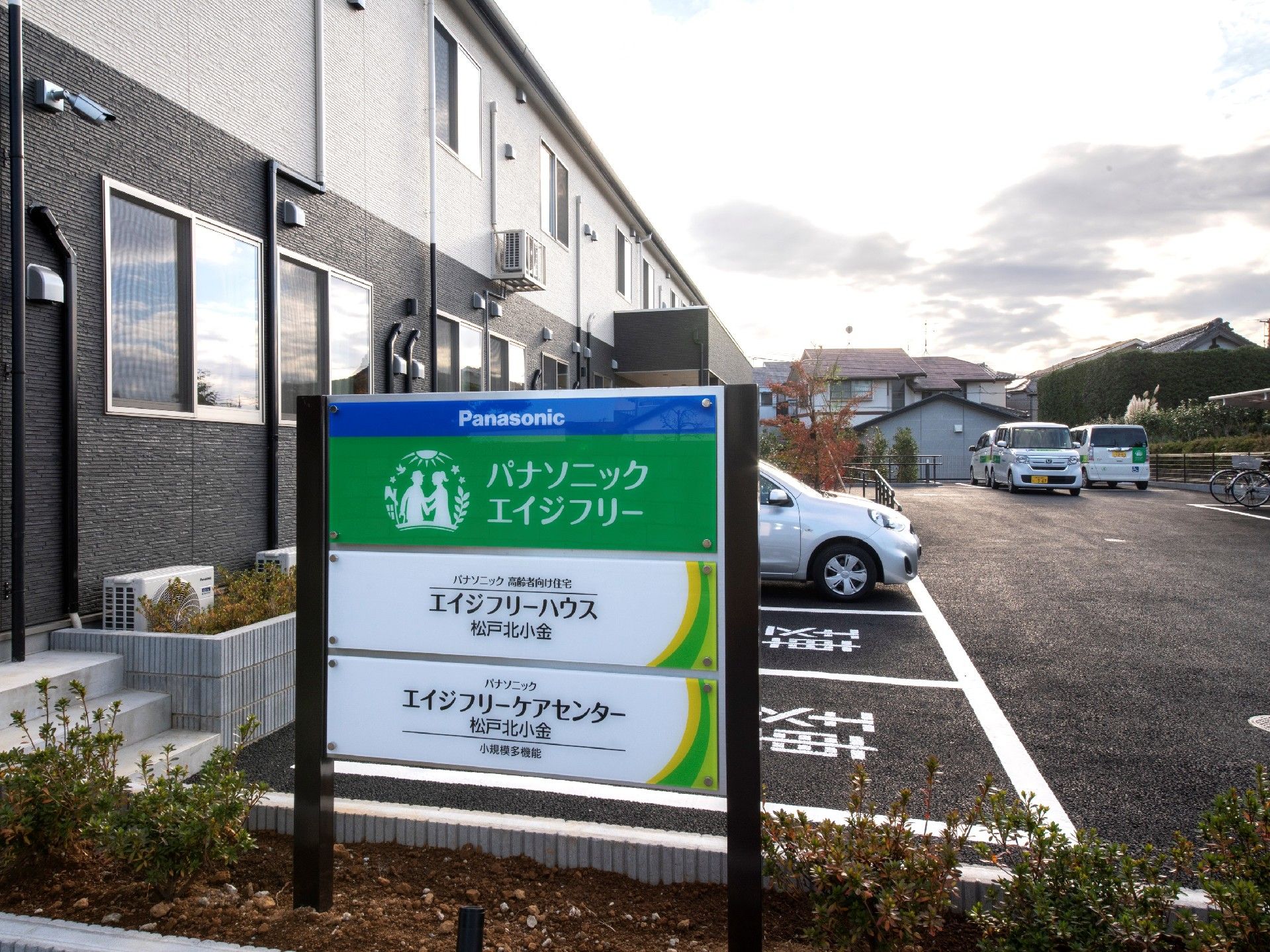 住宅と併設事業所の名前が書かれたプレートが貼られた看板の奥には駐車スペースと2階建ての建物がある。