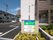 住宅や併設事業所の名前が書かれた門のそばには木が植えられ、駐車場や建物が奥に見える。