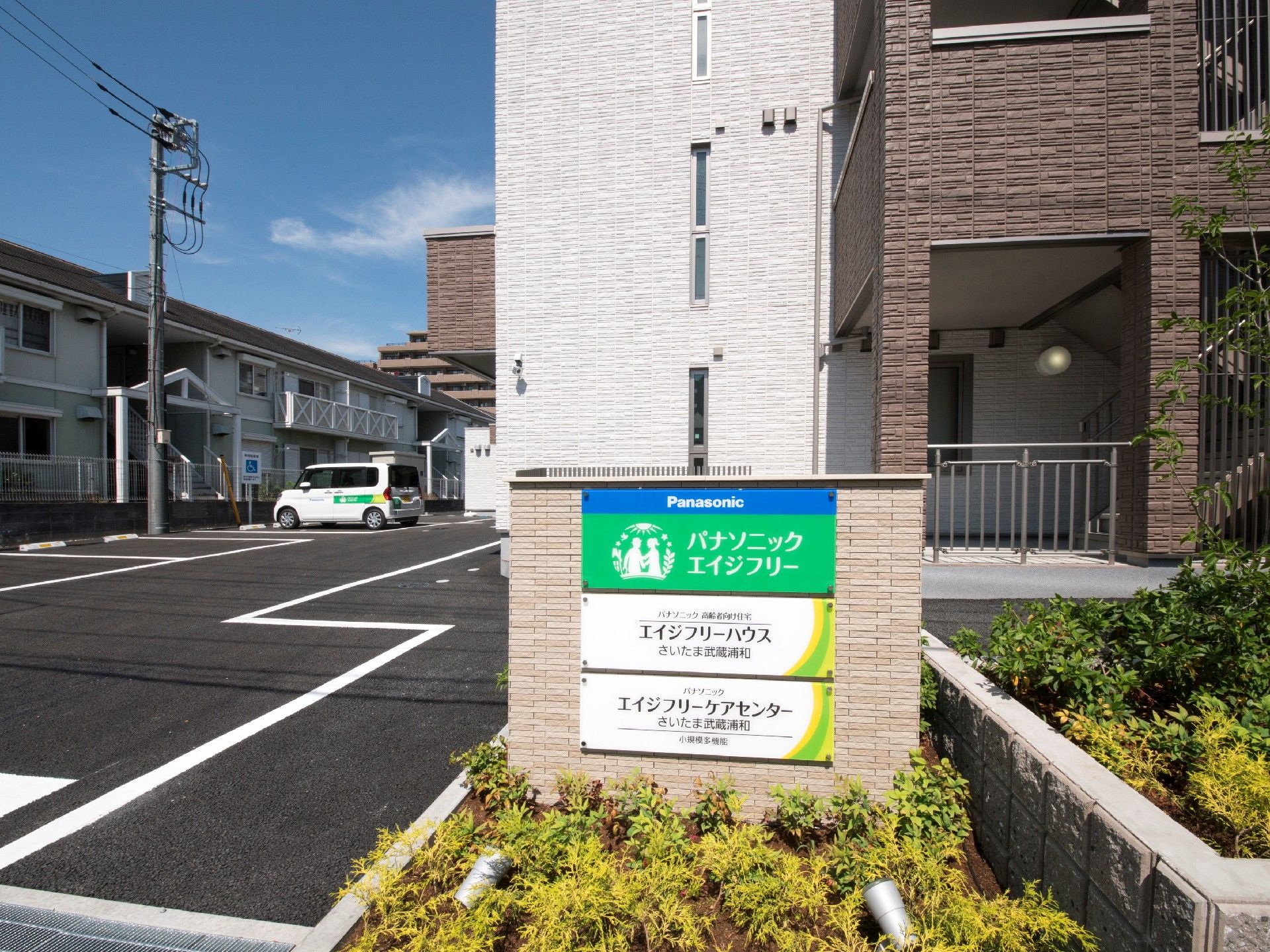 ブラウン系の建物の前には住宅や併設事業所の名前が書かれた門があり、広い駐車場もある。