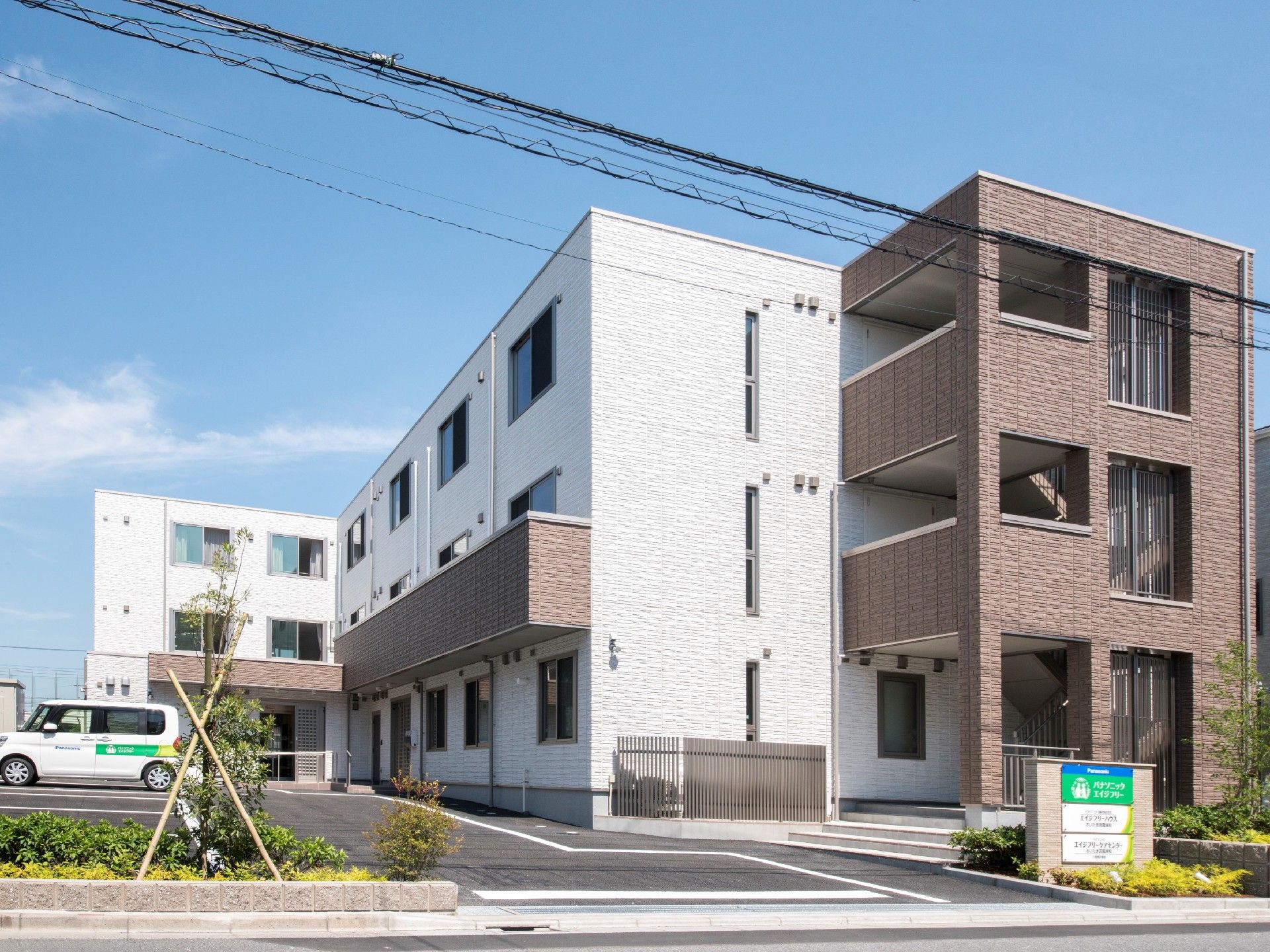 白とブラウンの3階建ての建物の周りには緑も多く、駐車場付き。玄関には屋根がある。