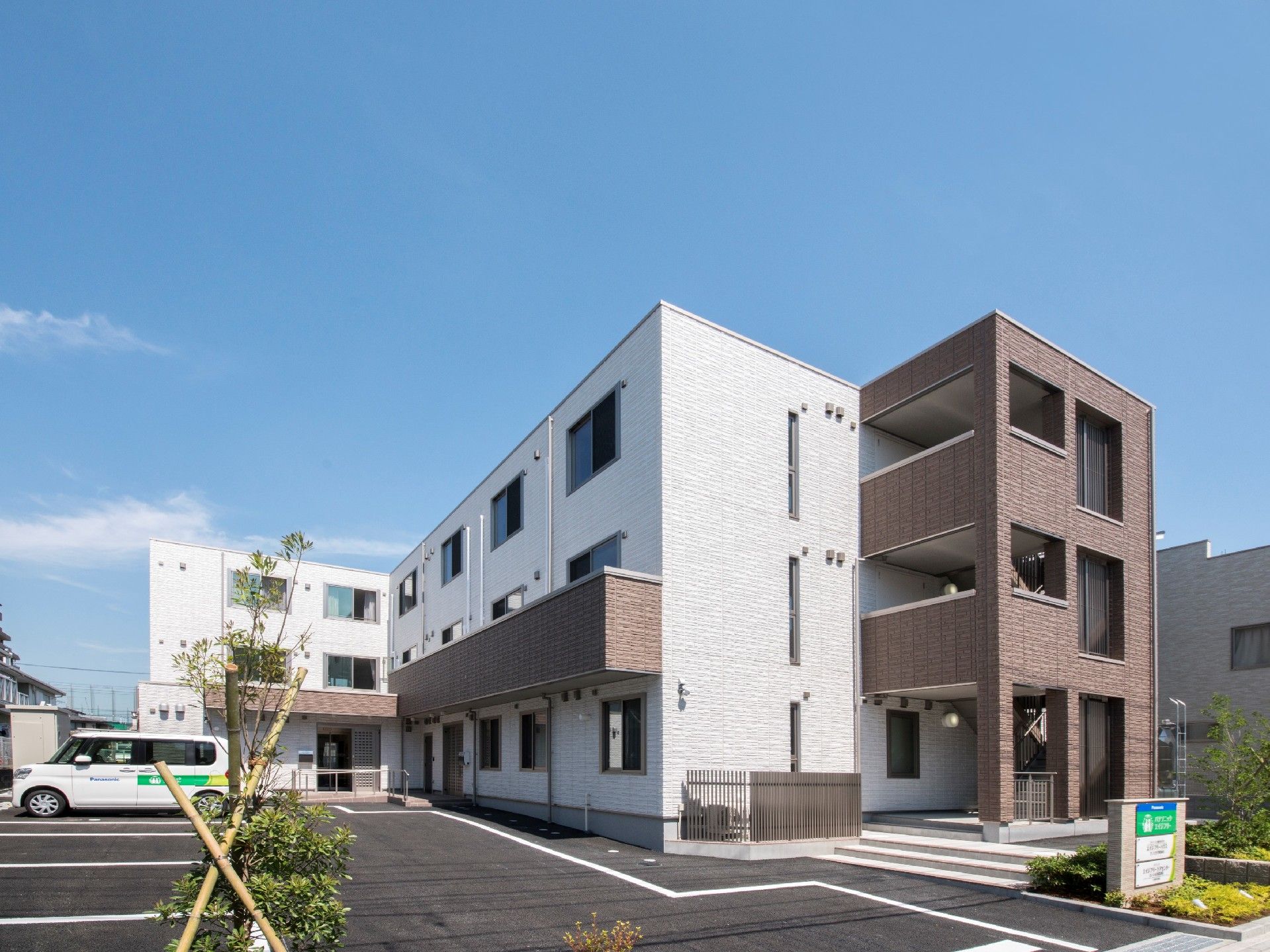 3階建ての建物はエル字型で屋外階段が付き、白い車が止まっている駐車場もある。