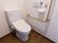 白い壁にフローリングの床のトイレに、洗浄機能付きの便座がある。壁面にＬ字型の手すり、便座のコントローラー、呼び出しボタンが設置されている。