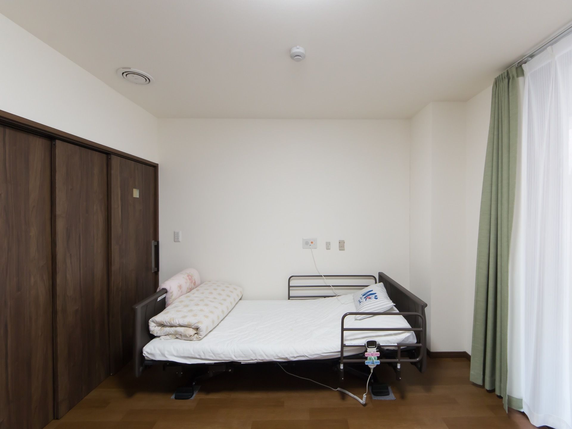 白い壁にフローリングの床の居室。扉は３連の引き戸で、カーテンの下がった大きな窓がある。ガードの付いた電動機能付きの大型ベッドがある。