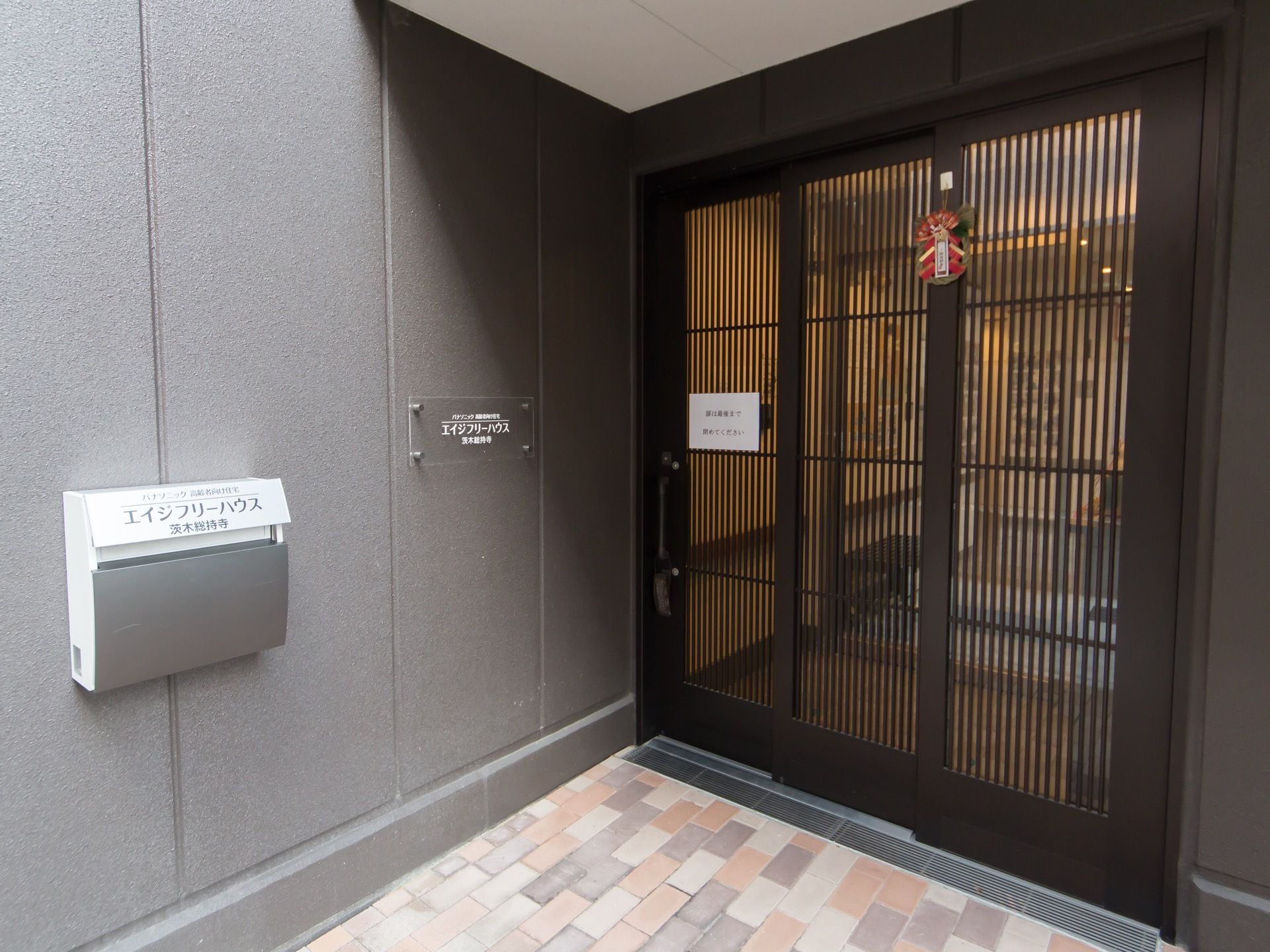 グレーの外壁にタイル張りの床になっている。３連の引き戸に正月飾りが付けられている。外壁には郵便受けと看板がかかっている。