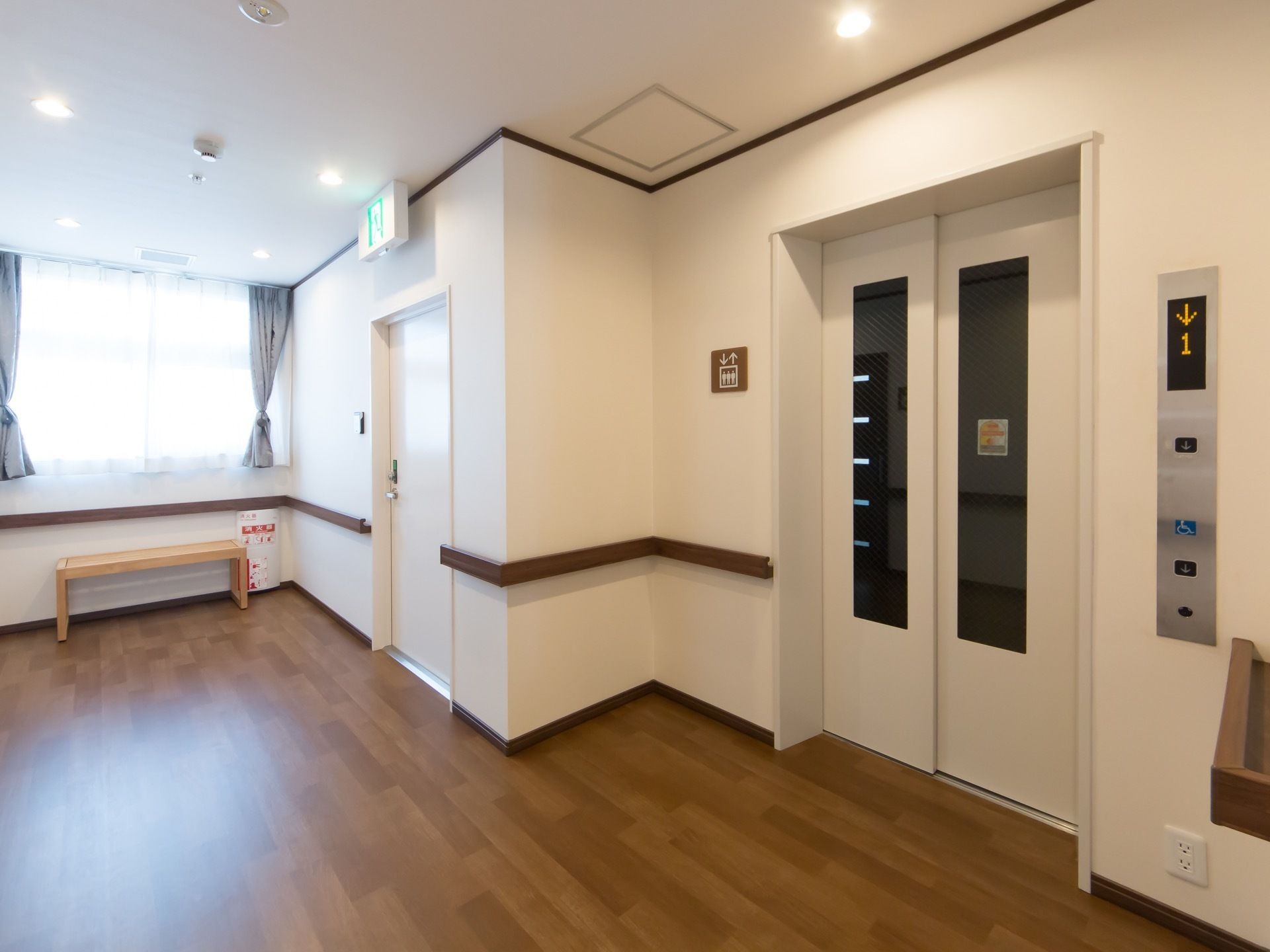 大きな窓付きのドアをもつエレベーターがある。乗り口の脇まで手すりが設けられている。左奥に窓が取られ、ベンチが置かれている。