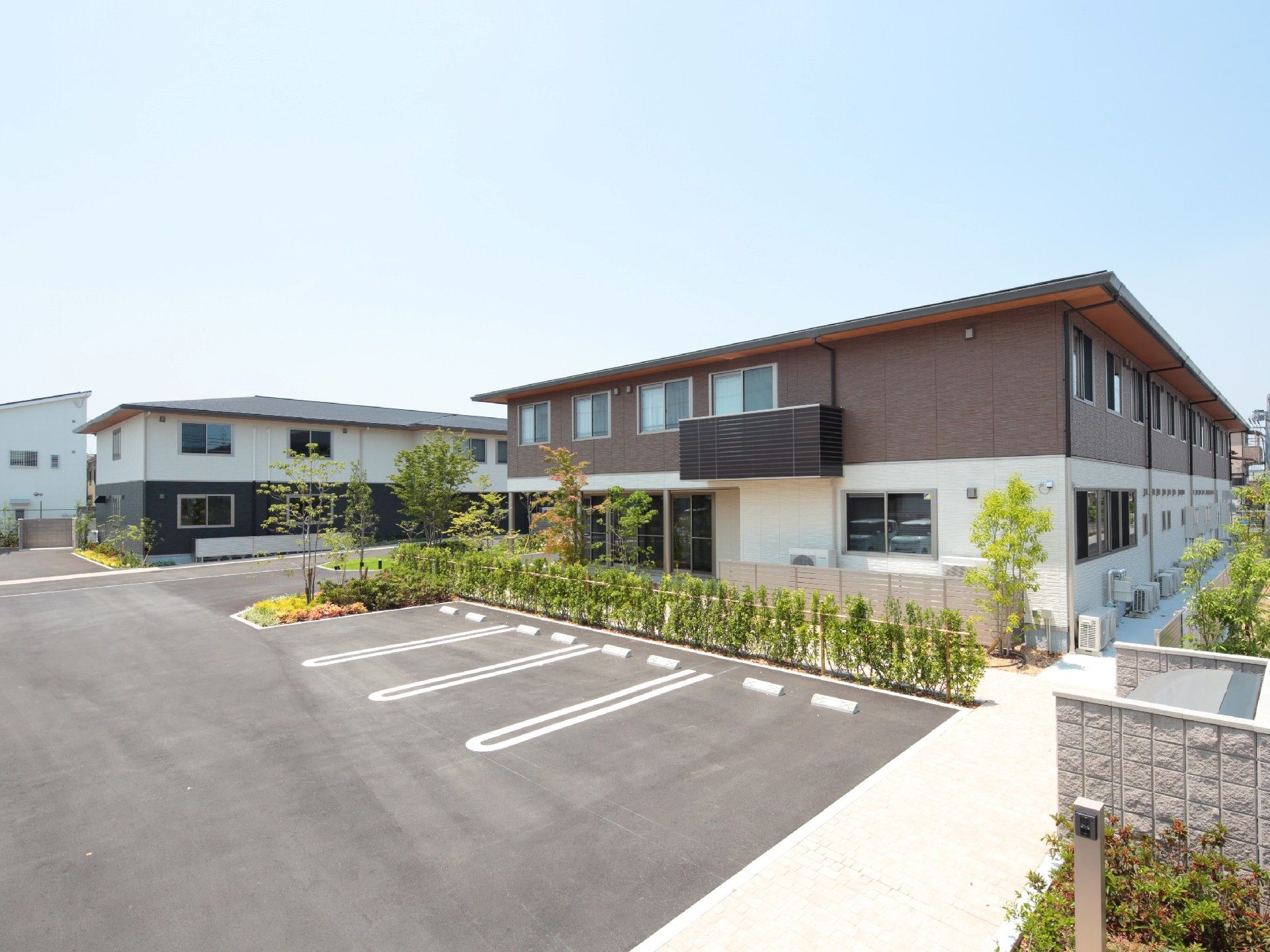 奥には白とダークグレーの2階建ての建物、手前にはブラウンと白の2階建ての建物があり、駐車場もある。