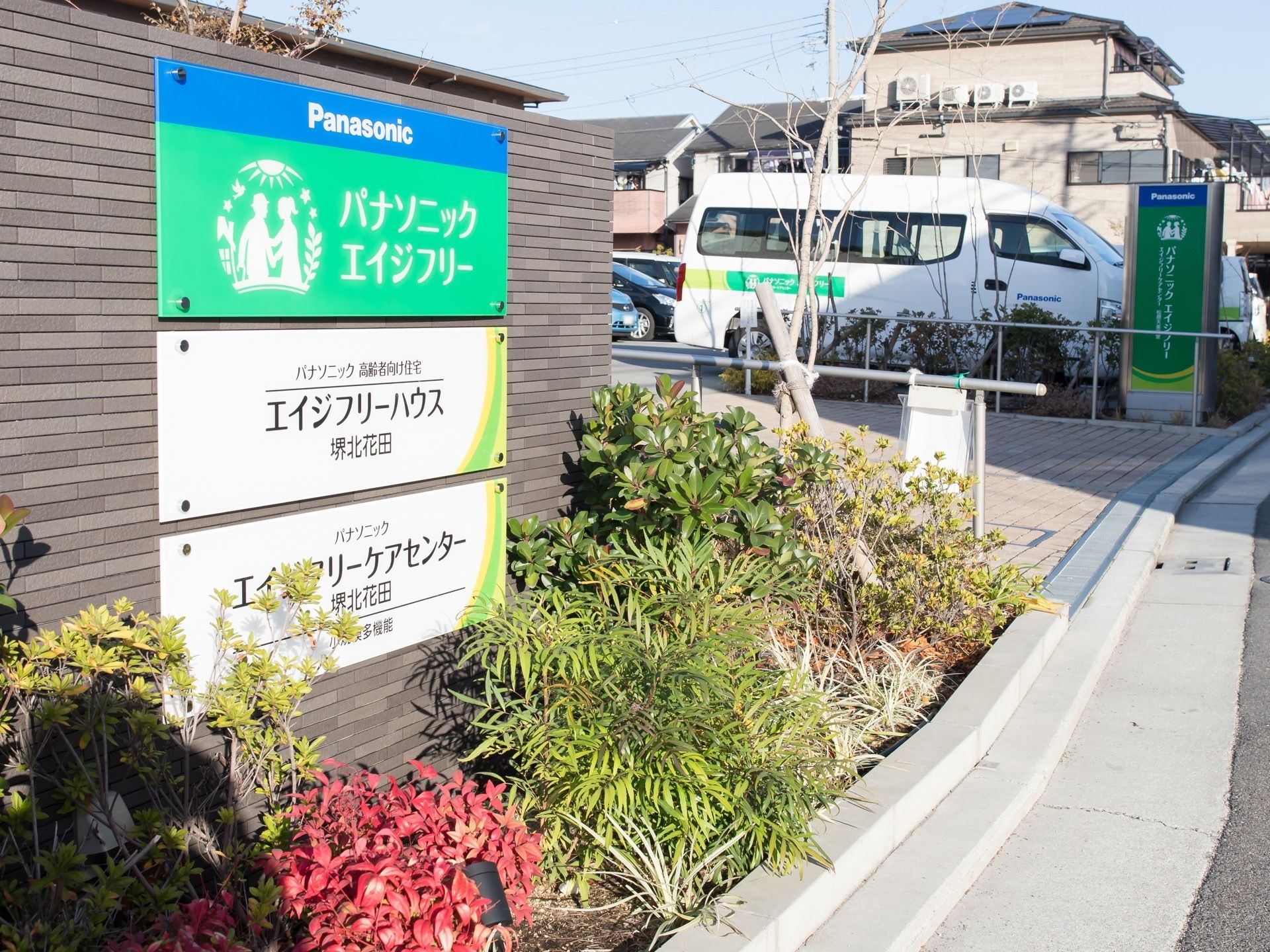 道路に面した敷地出入口にある塀に、３枚の看板が表示されている。一番上に運営会社の名称、下２枚に提供サービスと施設名称が書かれている。