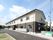 グレーと白の建物は２階建てで、門には住宅と併設事業所の看板が付き、駐車場も設置されている。