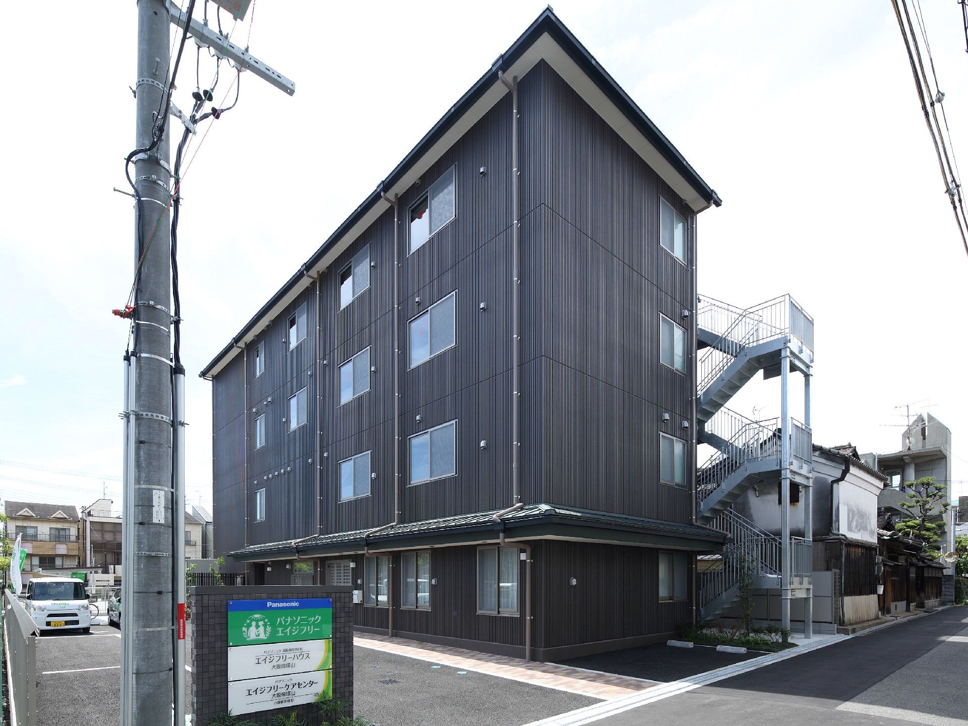 4階建ての建物。ダークグレーでまとめられ、看板や大きな駐車スペース、屋外階段が付いている。