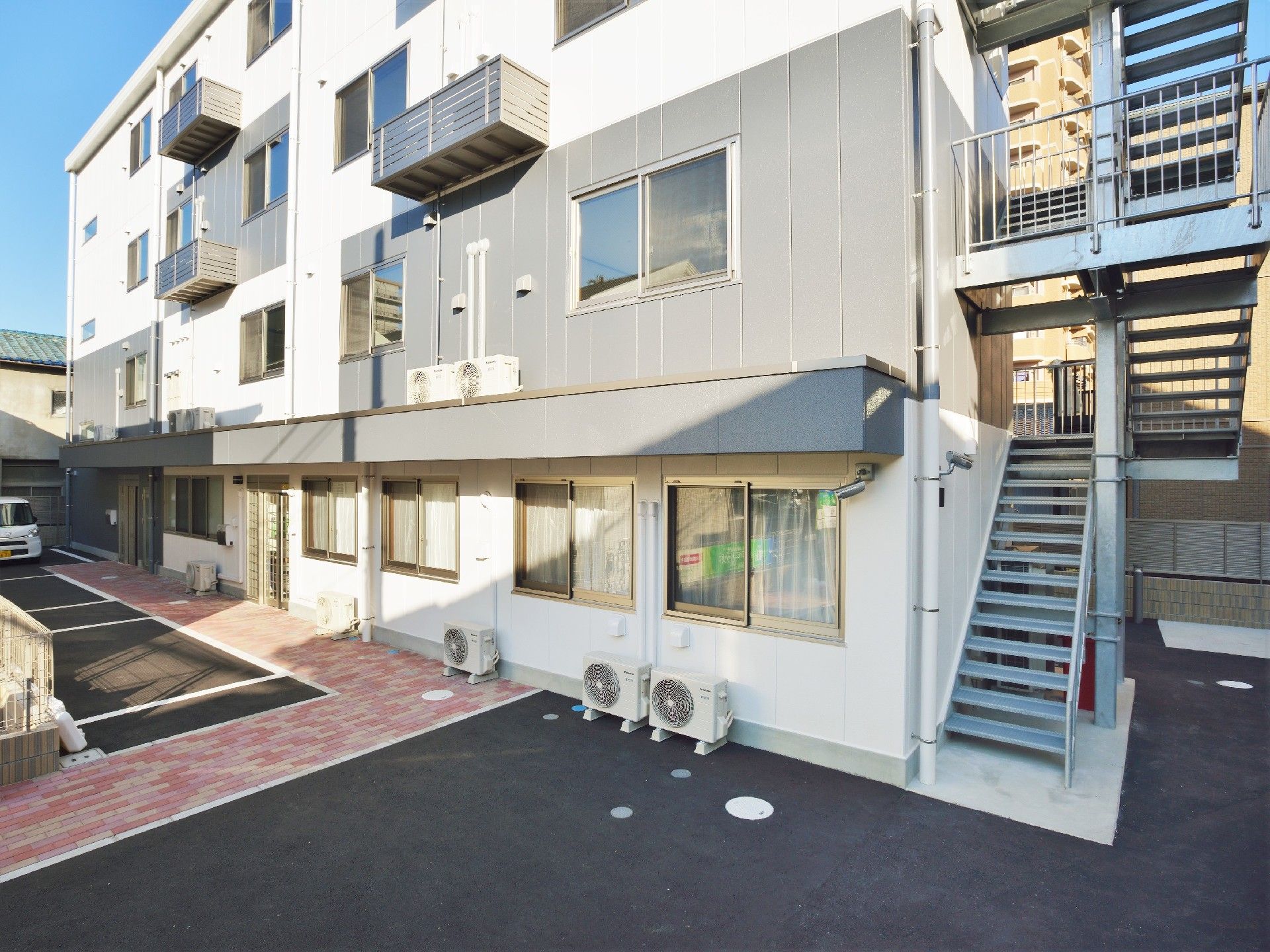 4階建ての建物は白とグレーで、側面には階段があり、駐車場も付いている。