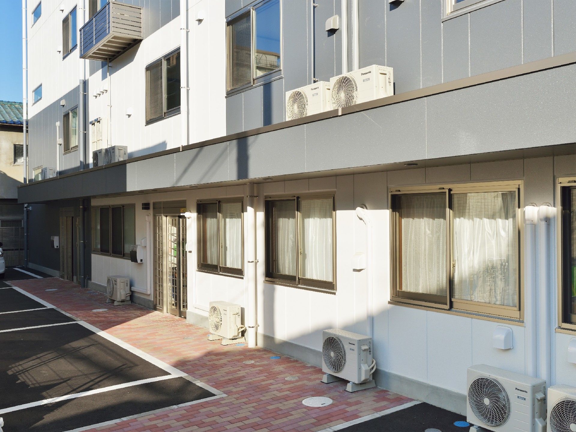 白とグレーの4階建ての建物の1階部分中央には出入り口が2か所あり、駐車スペースも用意されている。