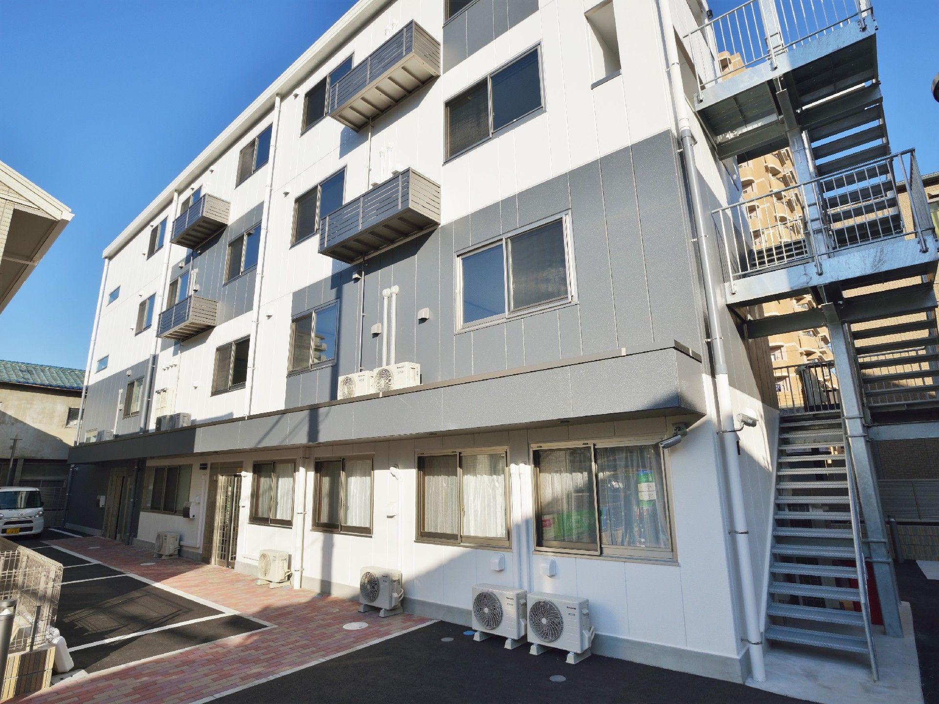 屋外階段が設置された白とグレーの4階建ての建物の外観。駐車スペースもある。