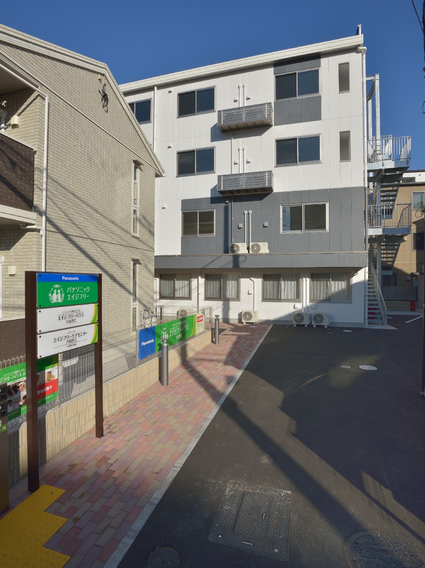 白とグレーの4階建ての建物の前にはレンガ調のアプローチが続いていて、周辺にも建物が多い。