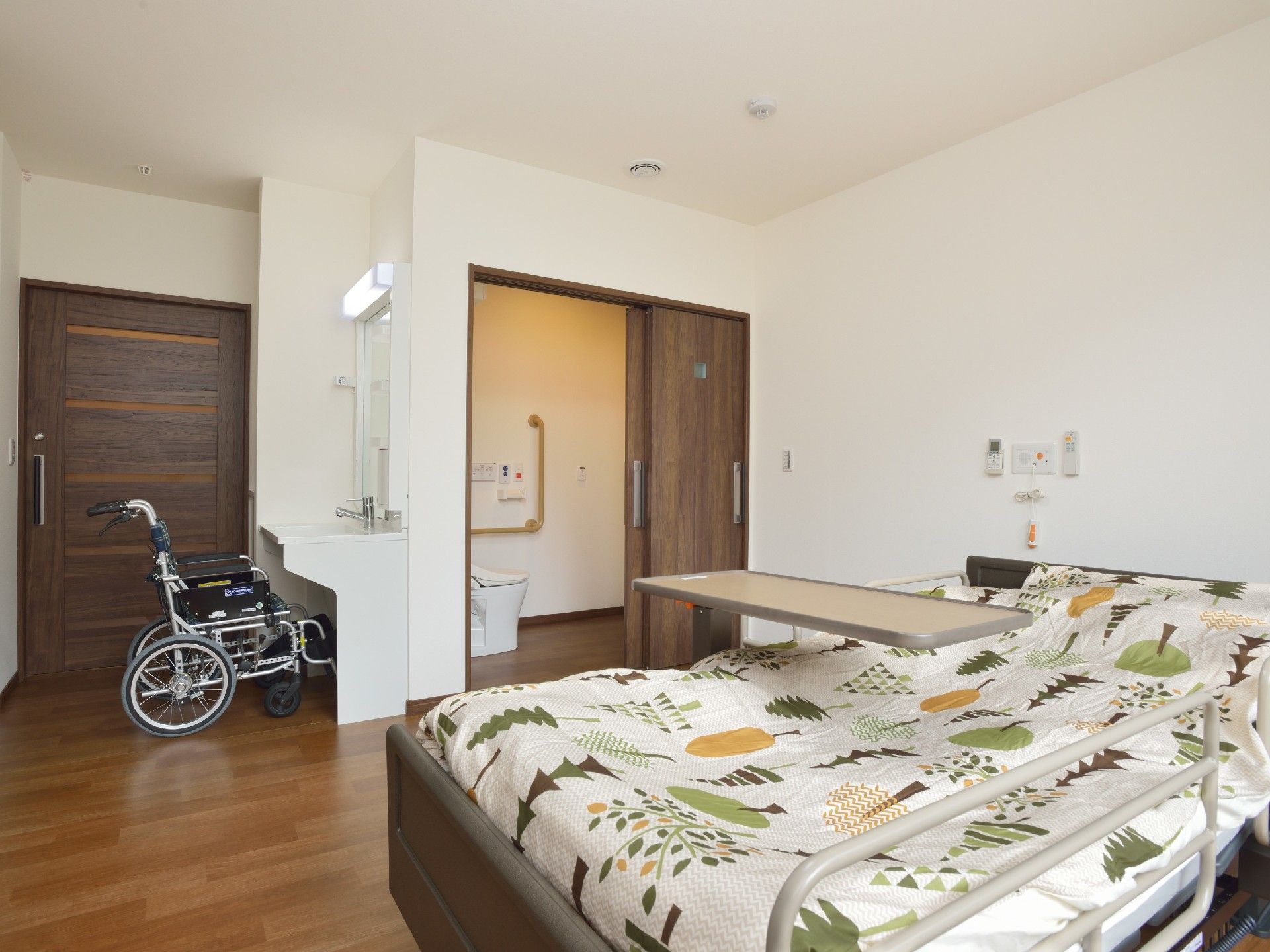 洗面所には車いすがセットされ、ベッドや車いす対応のトイレもある居室。ベッドにはテーブルも付いている。