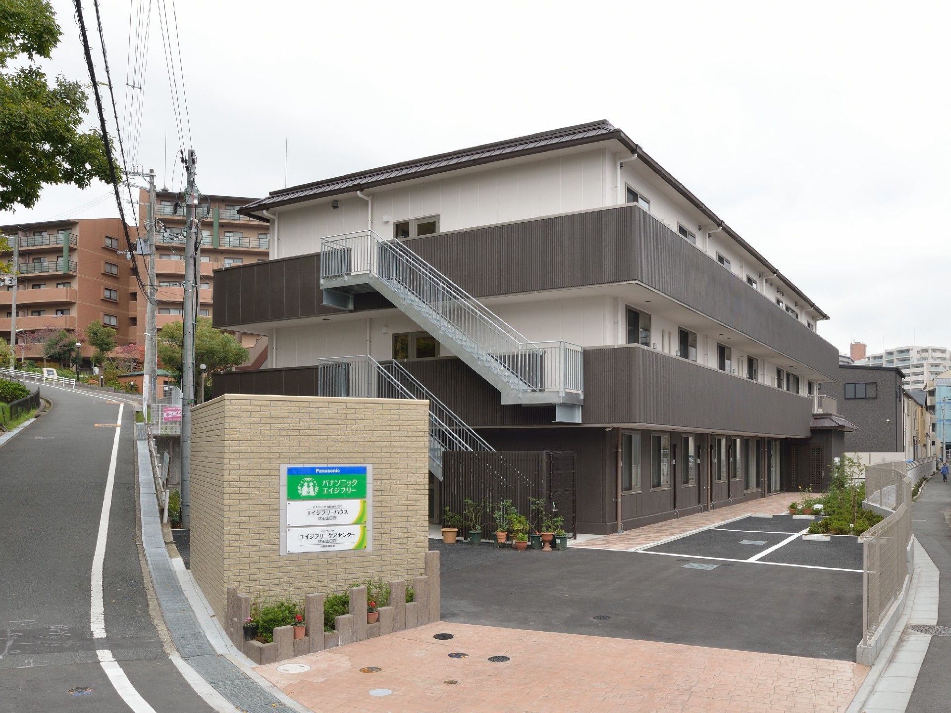住宅地に立つ3階建ての建物。白とダークグレーでまとめられ、敷地内には駐車場もある。