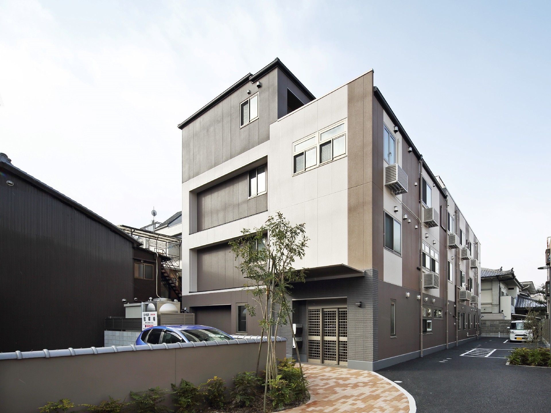 4階建ての建物。ダークグレーと白でまとめられ、大きな駐車スペースやアプローチが付いている。