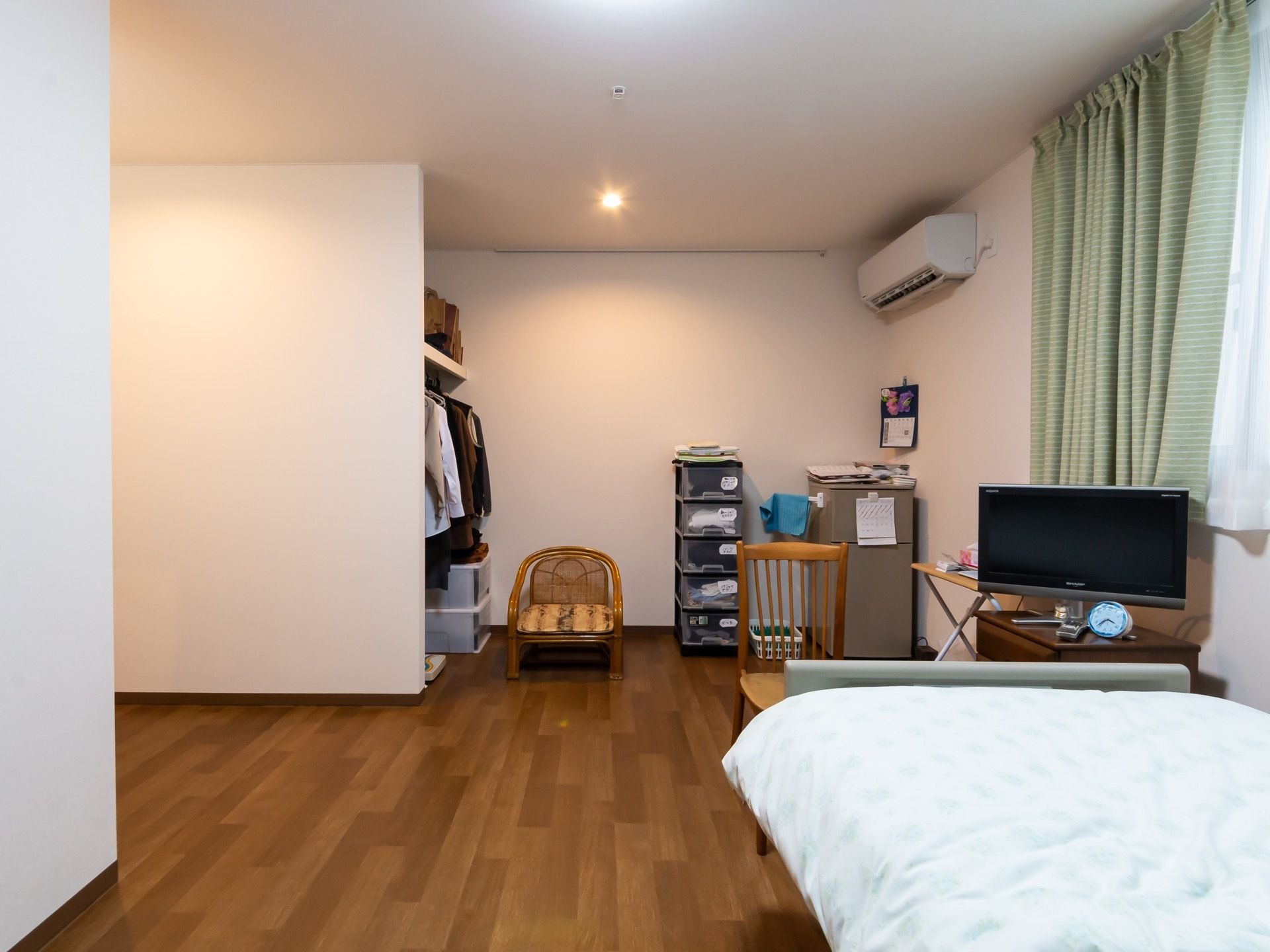 落ち着いた色合いの、フローリング部屋である。エアコンが備え付けられているほか、テレビやベッドも置かれている。