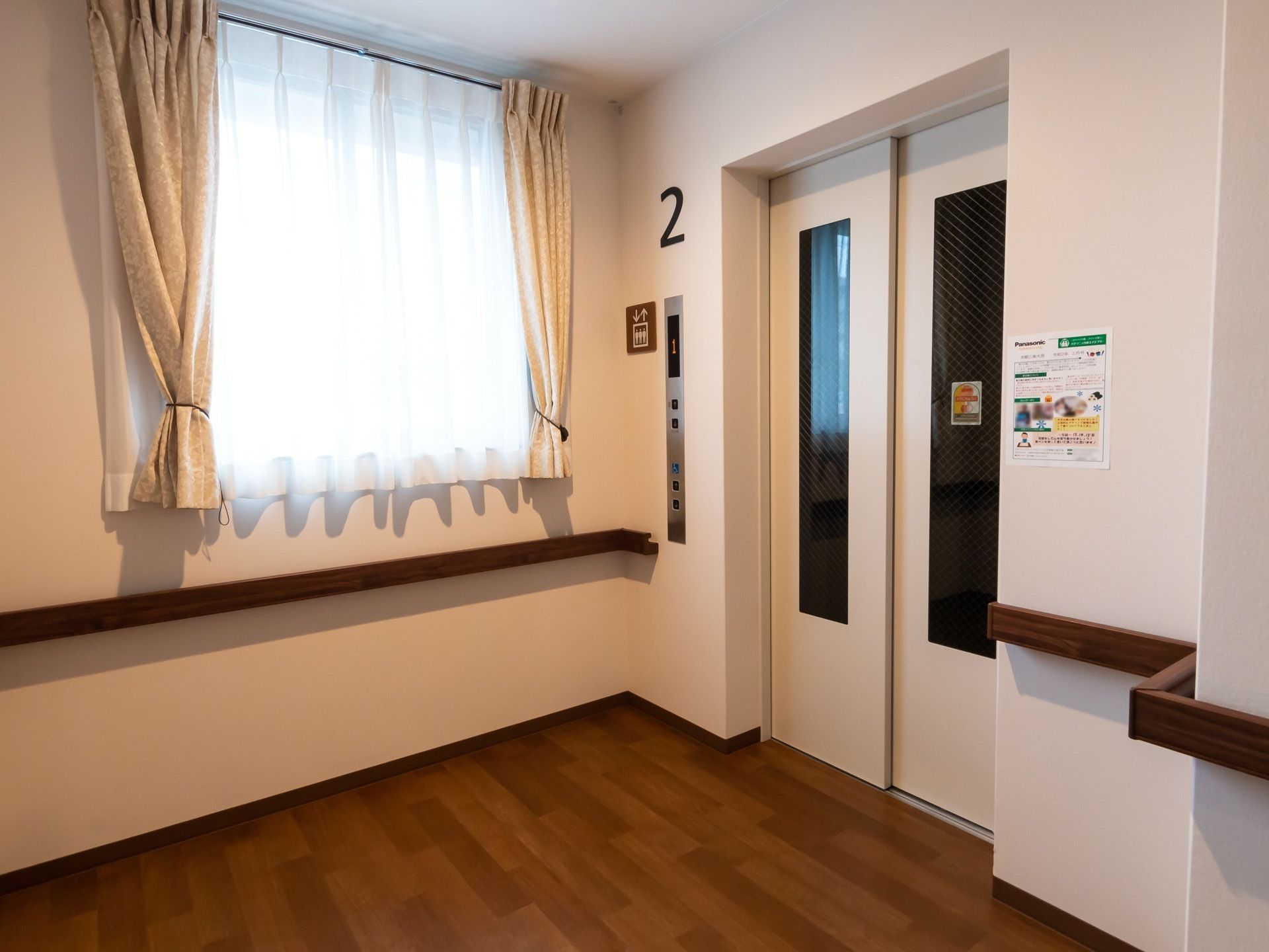 通路の突き当りにエレベーターがある。白色のスタイリッシュなデザイン。壁伝いには、木製の手すりが備え付けられている。