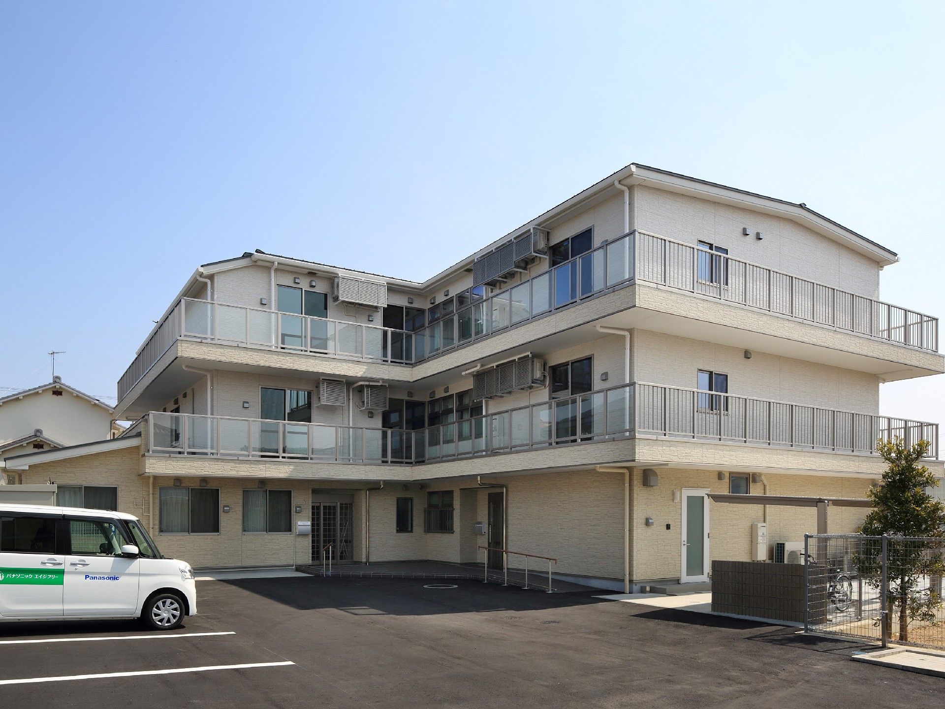3階建てのベージュの建物にはベランダが付き、玄関の周りには木も植えられている。