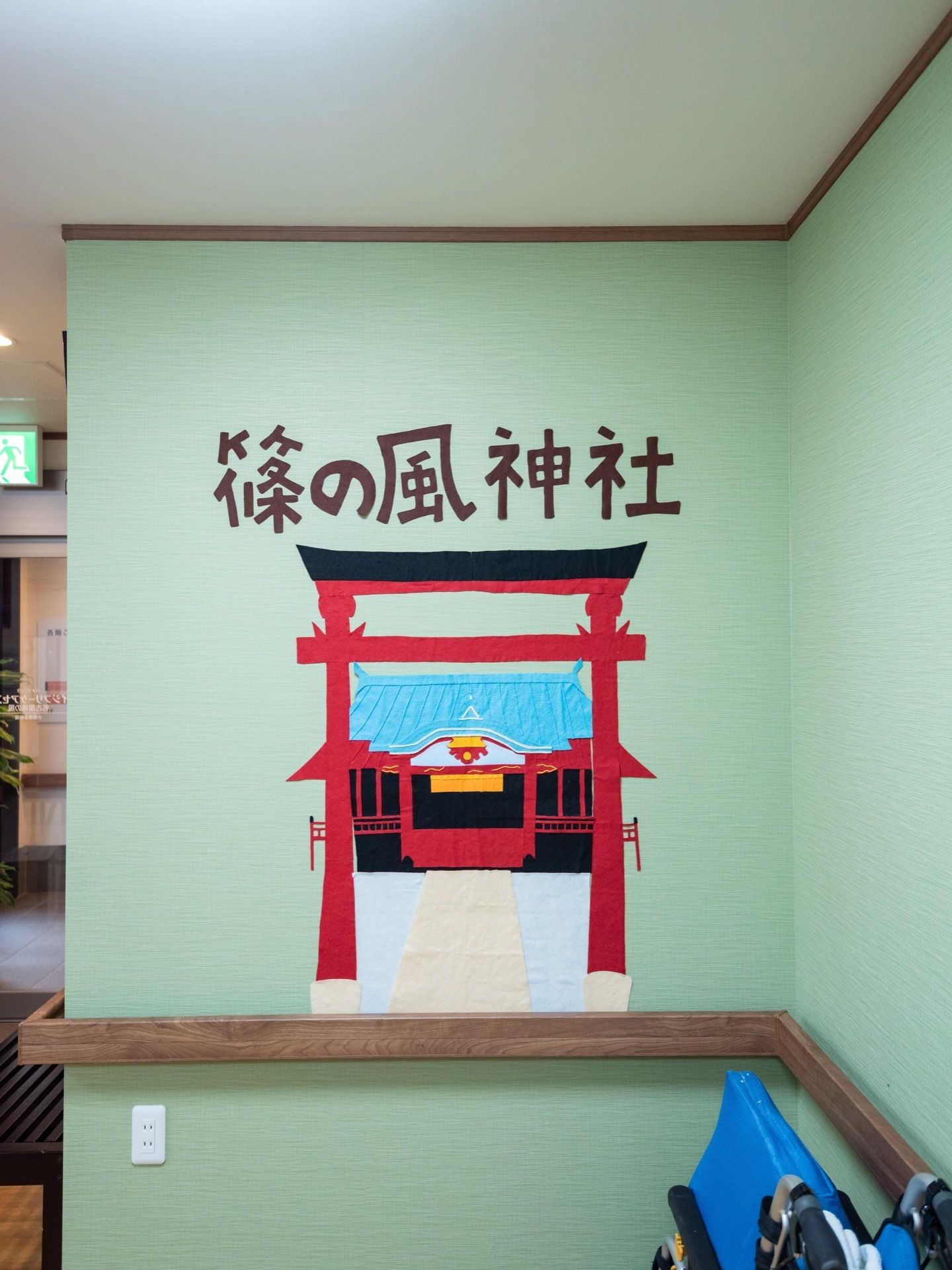 薄緑の壁に、施設名を冠した神社名と神殿、鳥居の絵が描かれている。下には、壁の周囲に設置された手すりと車椅子がある。
