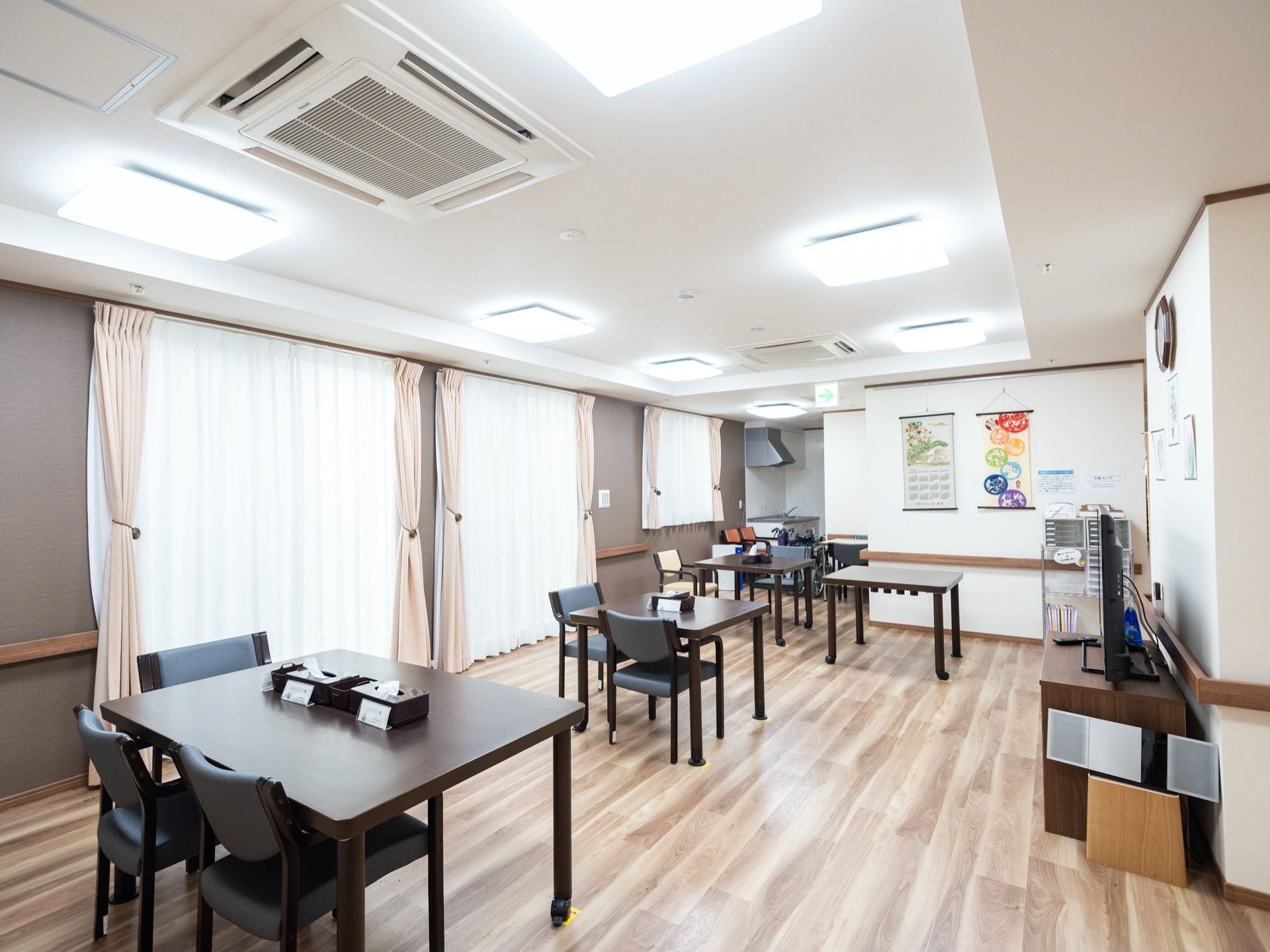 焦げ茶色の正方形のテーブルが複数置かれている。その周りには青色の背もたれのある椅子がある。壁には時計がある。