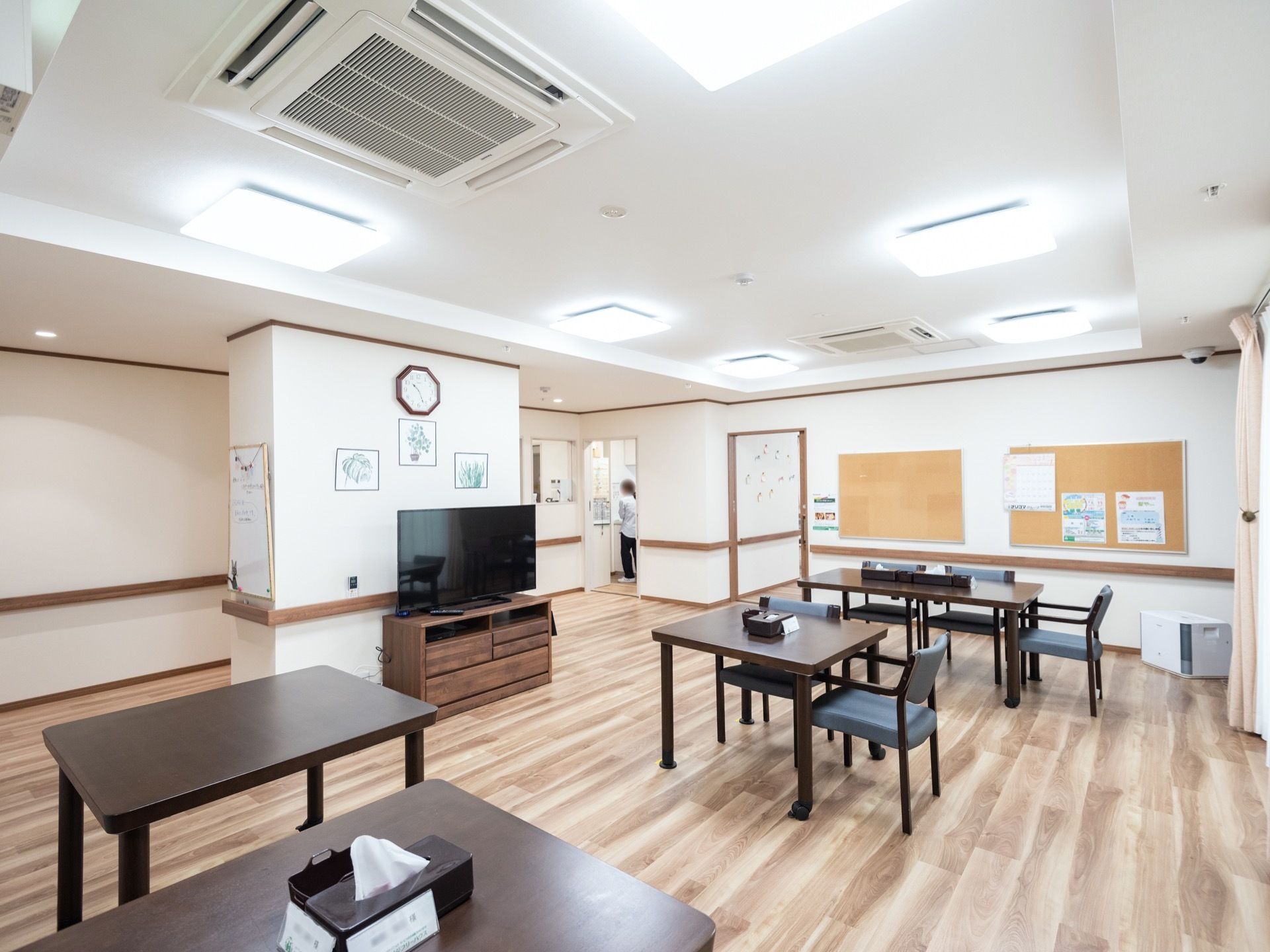 天井には、規則正しく照明器具が配置され、エアコンが埋め込まれている。壁際にはテレビが１台設置されており、テーブルと椅子が複数置かれている。