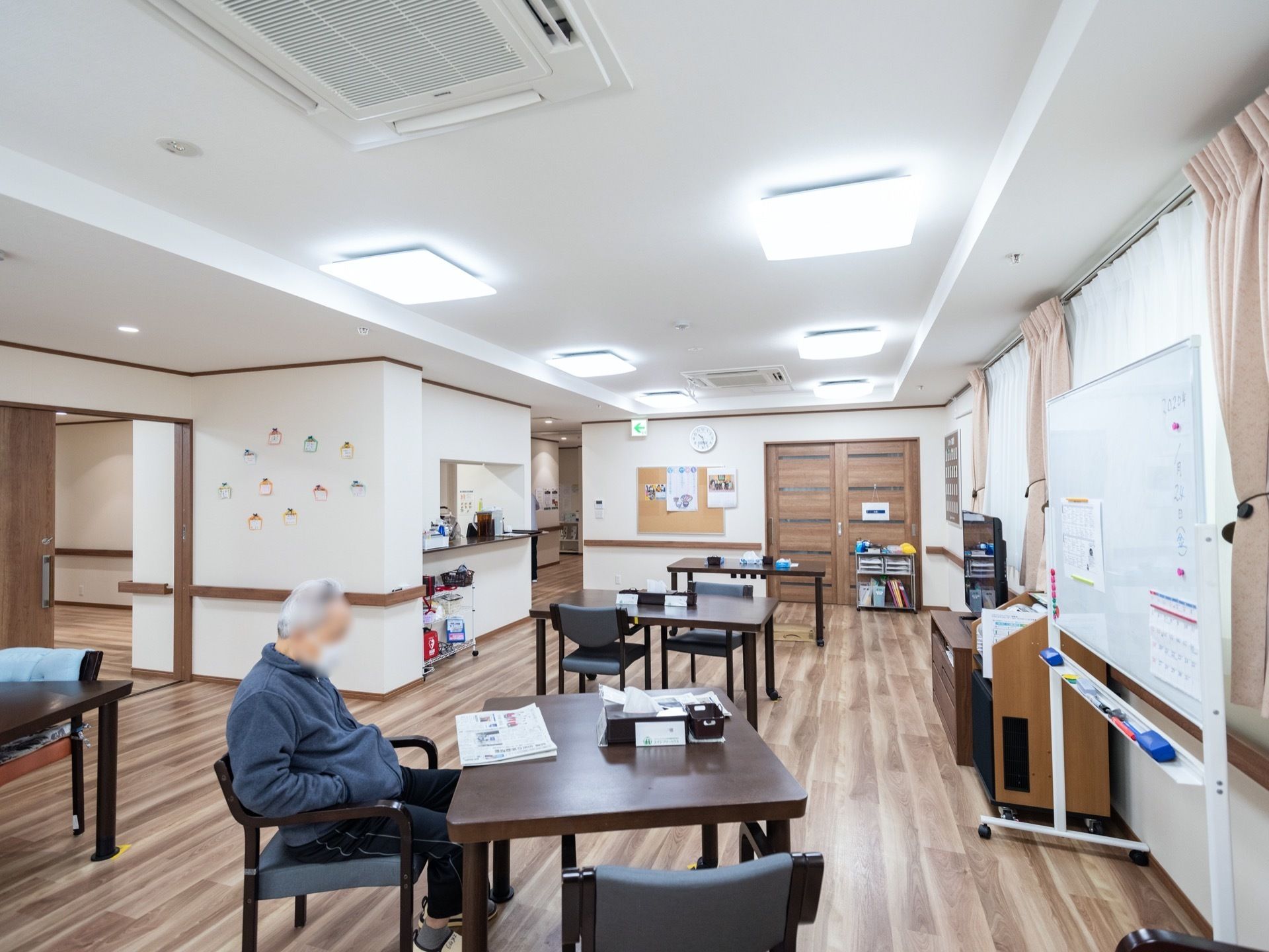 複数のテーブルと椅子が置かれており、１人の入居者様が椅子に座り、新聞を読んでいる様子である。壁には、飾りつけがされている。
